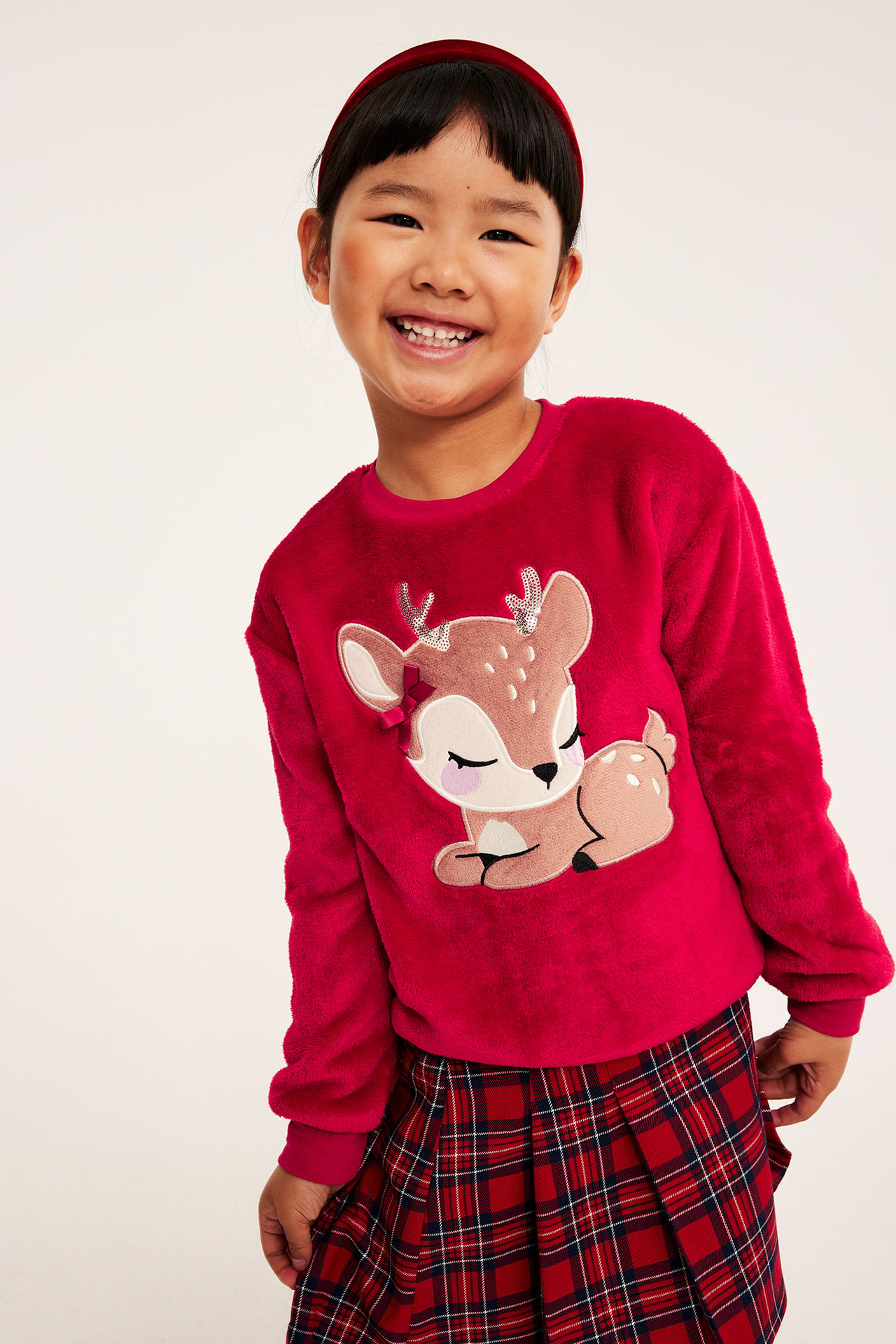 Frontvy på barn i fuchsia fluffig topp med rådjursapplikation, rödrutig kjol och rött hårband.