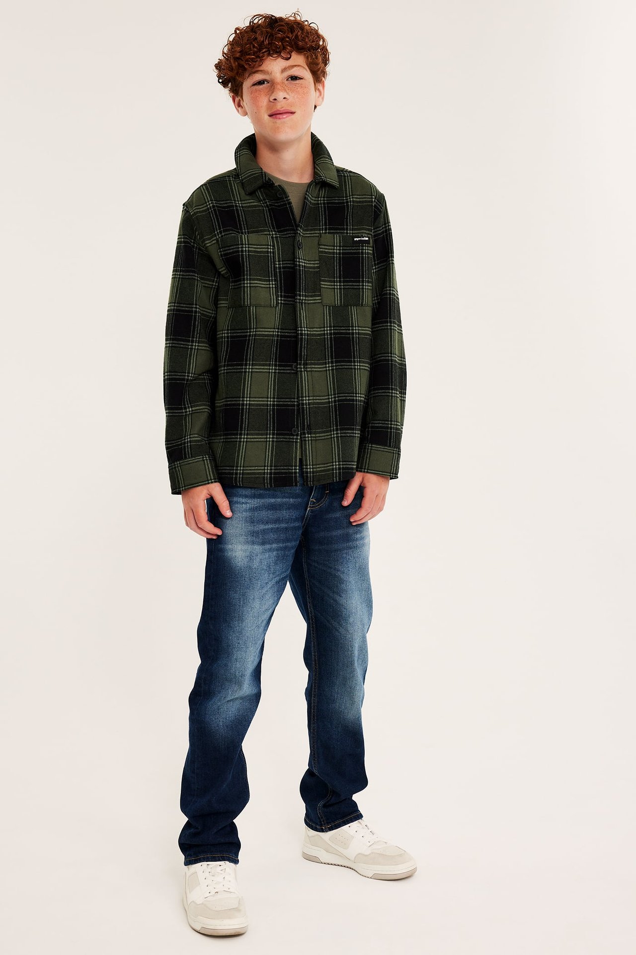 Framifrån, barn i grön- och svartrutig skjorta över t-shirt, blå jeans och vita sneakers.