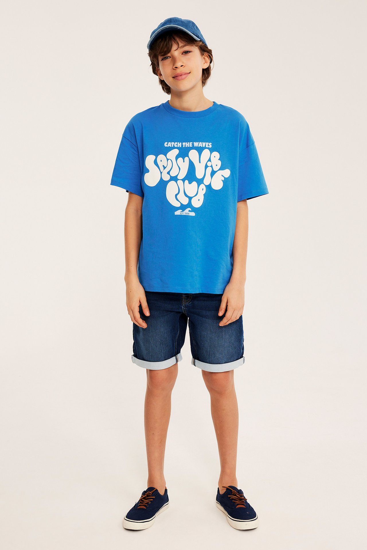 Etunäkymä: Lapsi sinisessä ylisuuressa t-paidassa etupainatuksella, tummissa denimshortseissa, sinisissä lenkkareissa ja farkkumyssassa.