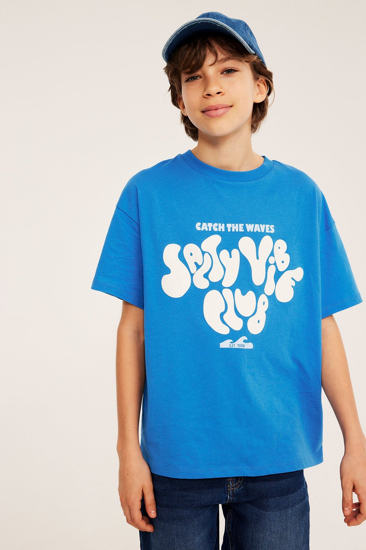 Framifrån, barn i blå t-shirt med vitt 'Salty Vibe Club'-tryck, stylad med blå jeanskeps och jeans.