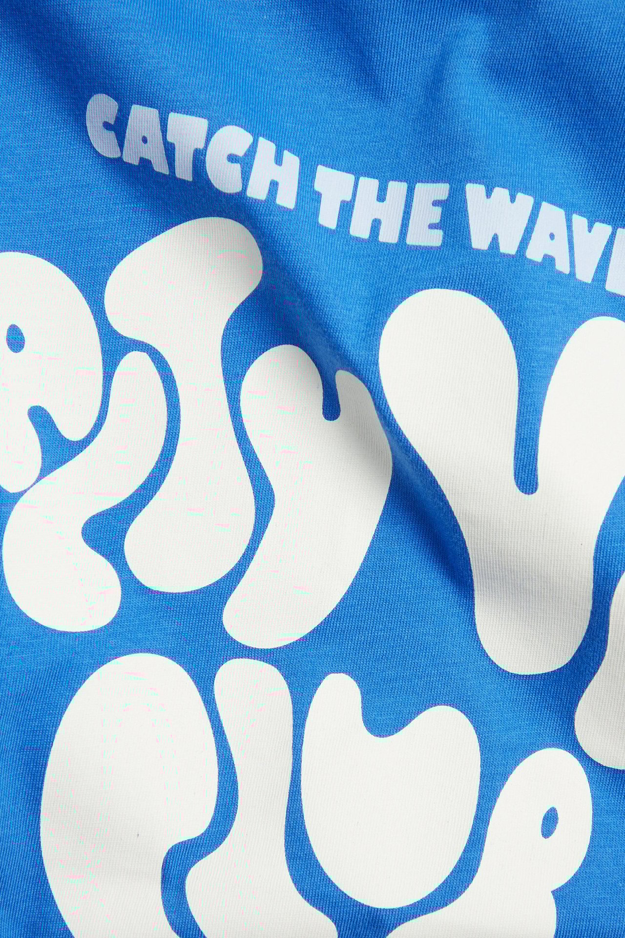 Zbliżenie niebieskiego, bawełnianego t-shirtu oversize z białym napisem 'CATCH THE WAVE' i abstrakcyjnym nadrukiem.