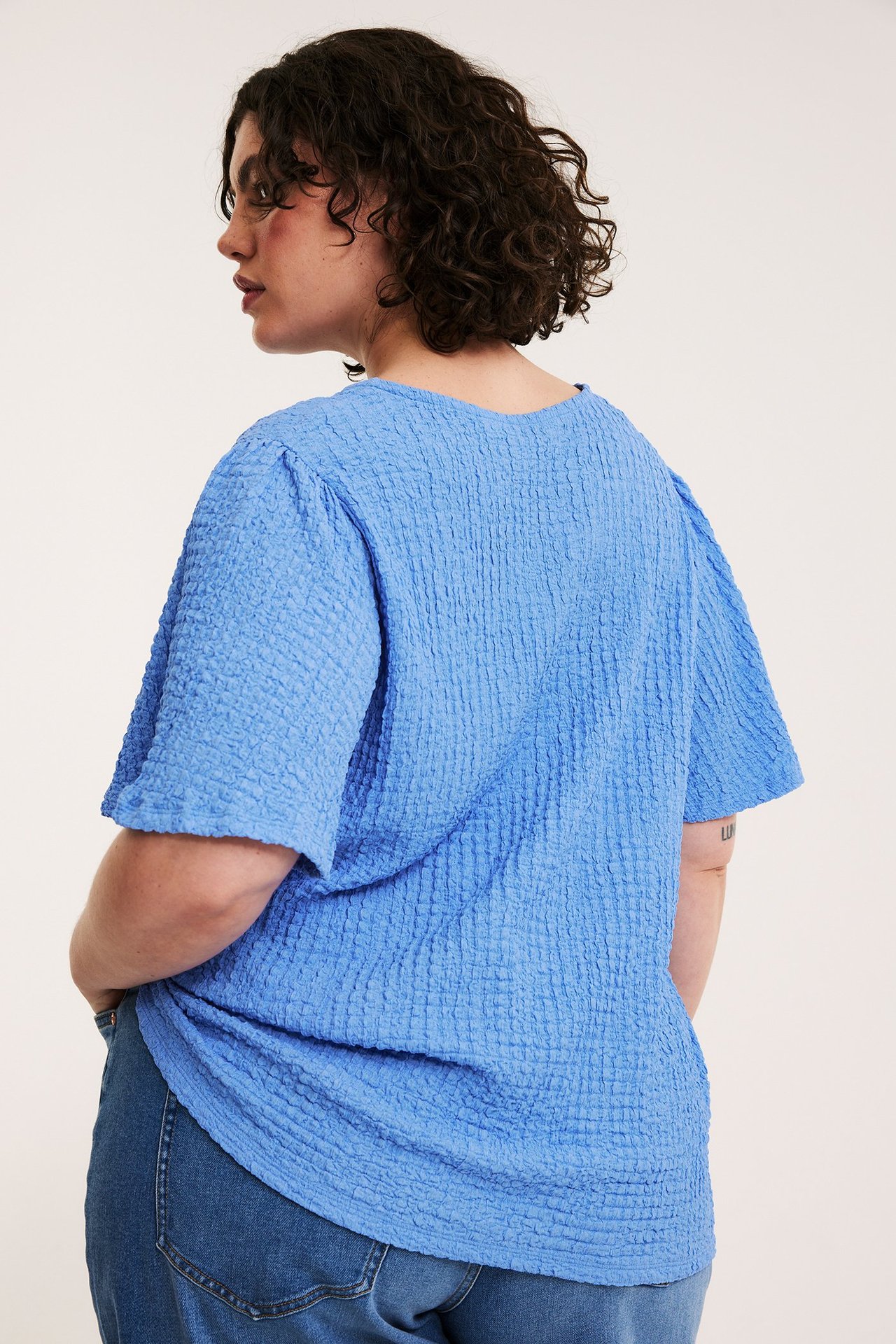 Nainen takaapäin sinisessä tekstuuripintaisessa lyhythihaisessa topissa ja sinisissä farkuissa.