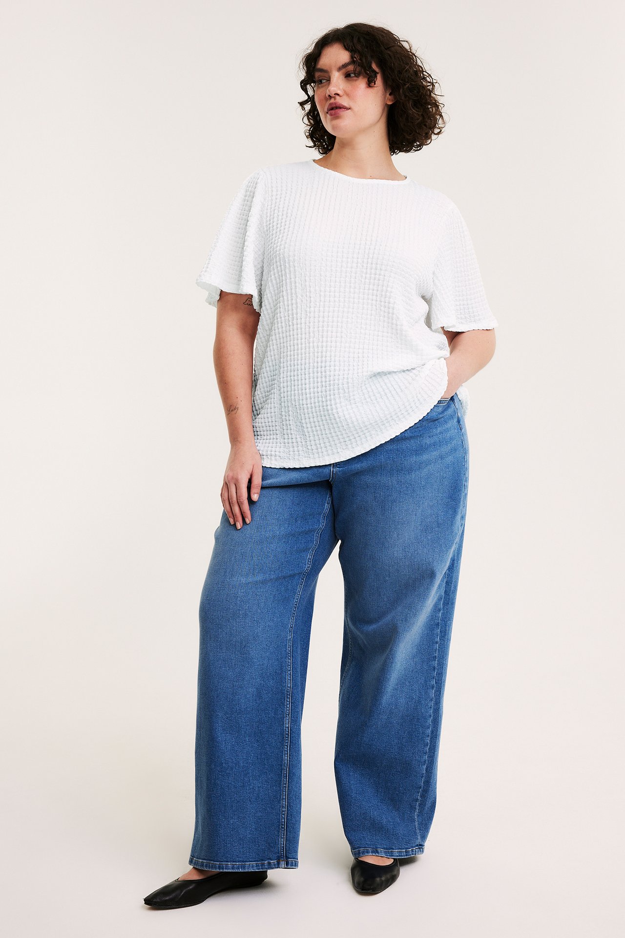 Naisen etukuva valkoisessa strukturoidussa lyhythihaisessa topissa, sinisissä leveälahkeisissa farkuissa ja mustissa kengissä.