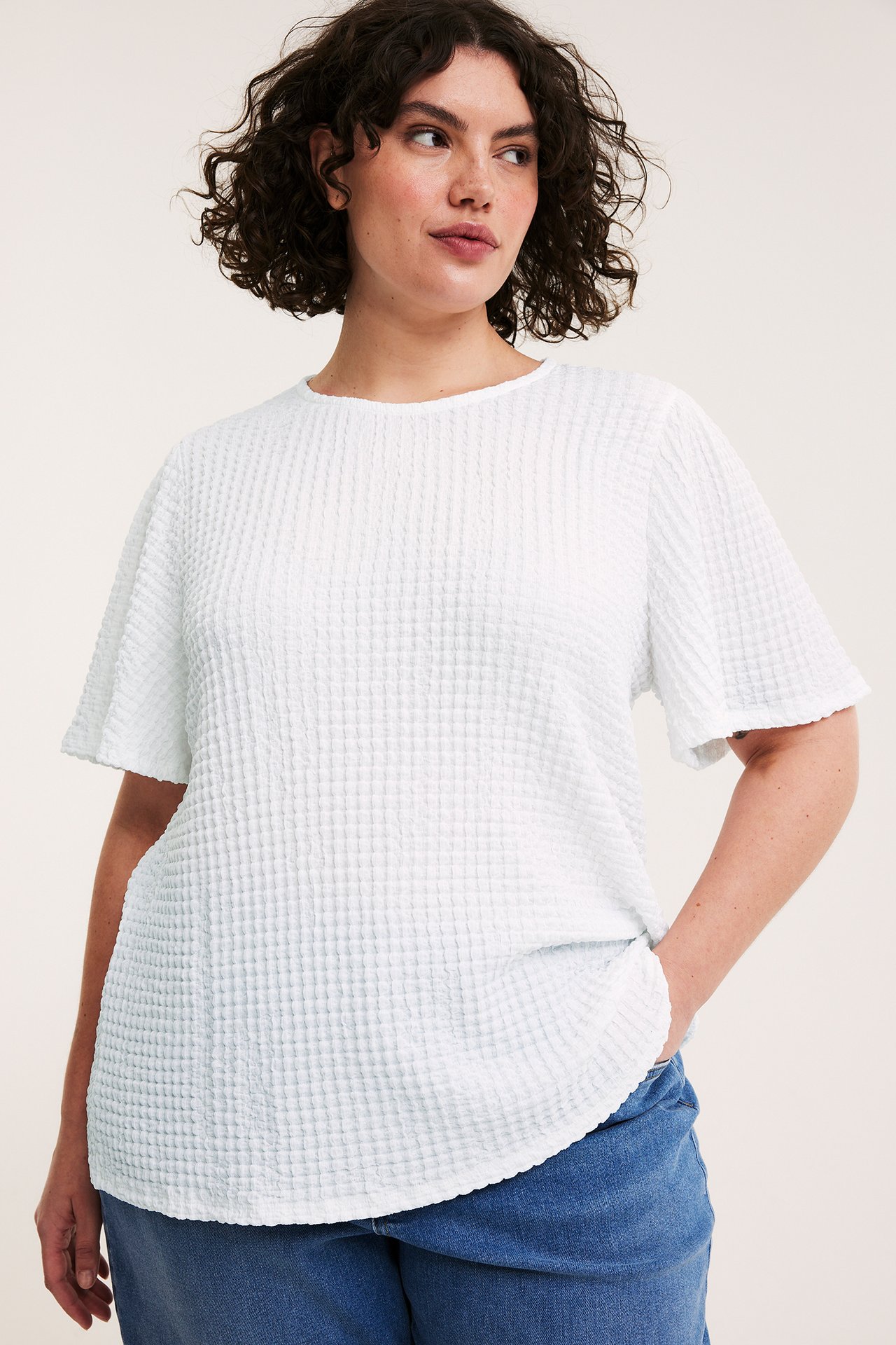 Widok z przodu: kobieta w białym, teksturowanym topie z krótkim rękawem i niebieskich dżinsach.