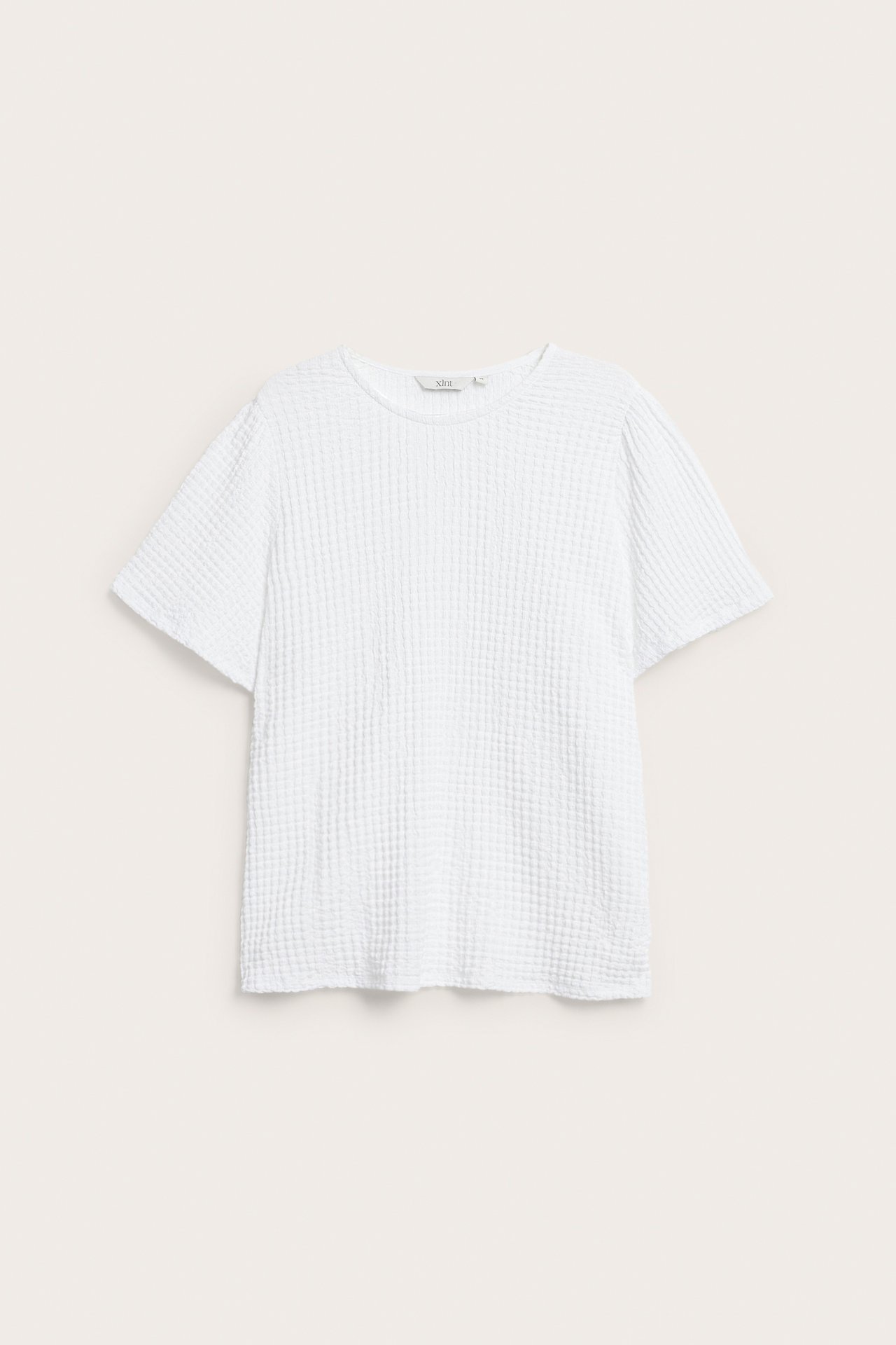 Edestä näkymä valkoisesta, väljästä, lyhythihaisesta topista, jossa on kuvioitu kangas ja pyöreä pääntie.