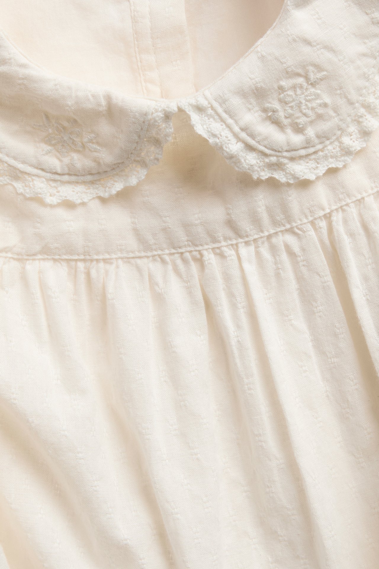 Nærbilde av en offwhite topp med brodert Peter Pan-krage og rynkede detaljer.