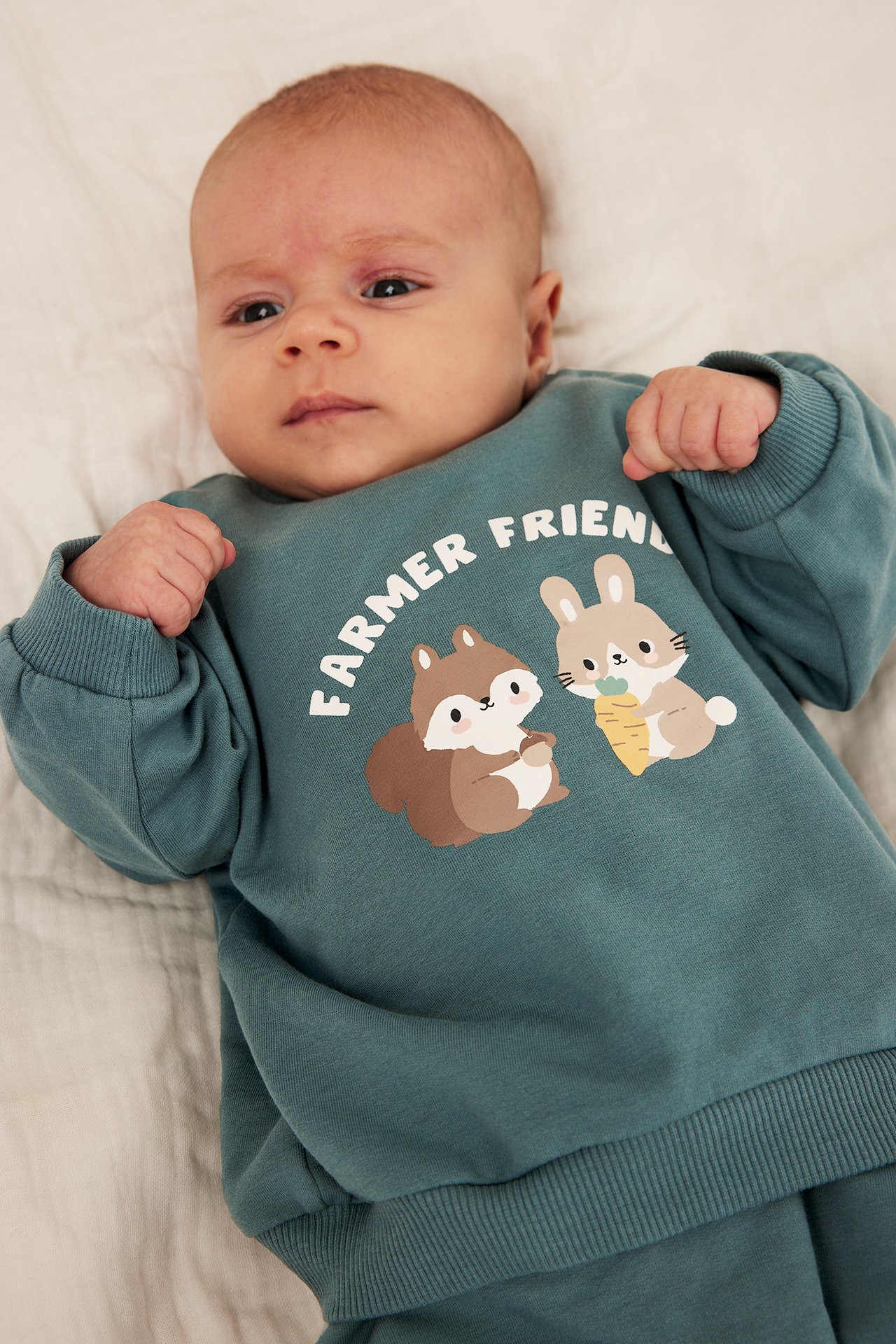 Framifrån: Bebis i turkos sweatshirt med djurtryck och texten 'FARMER FRIEND'.