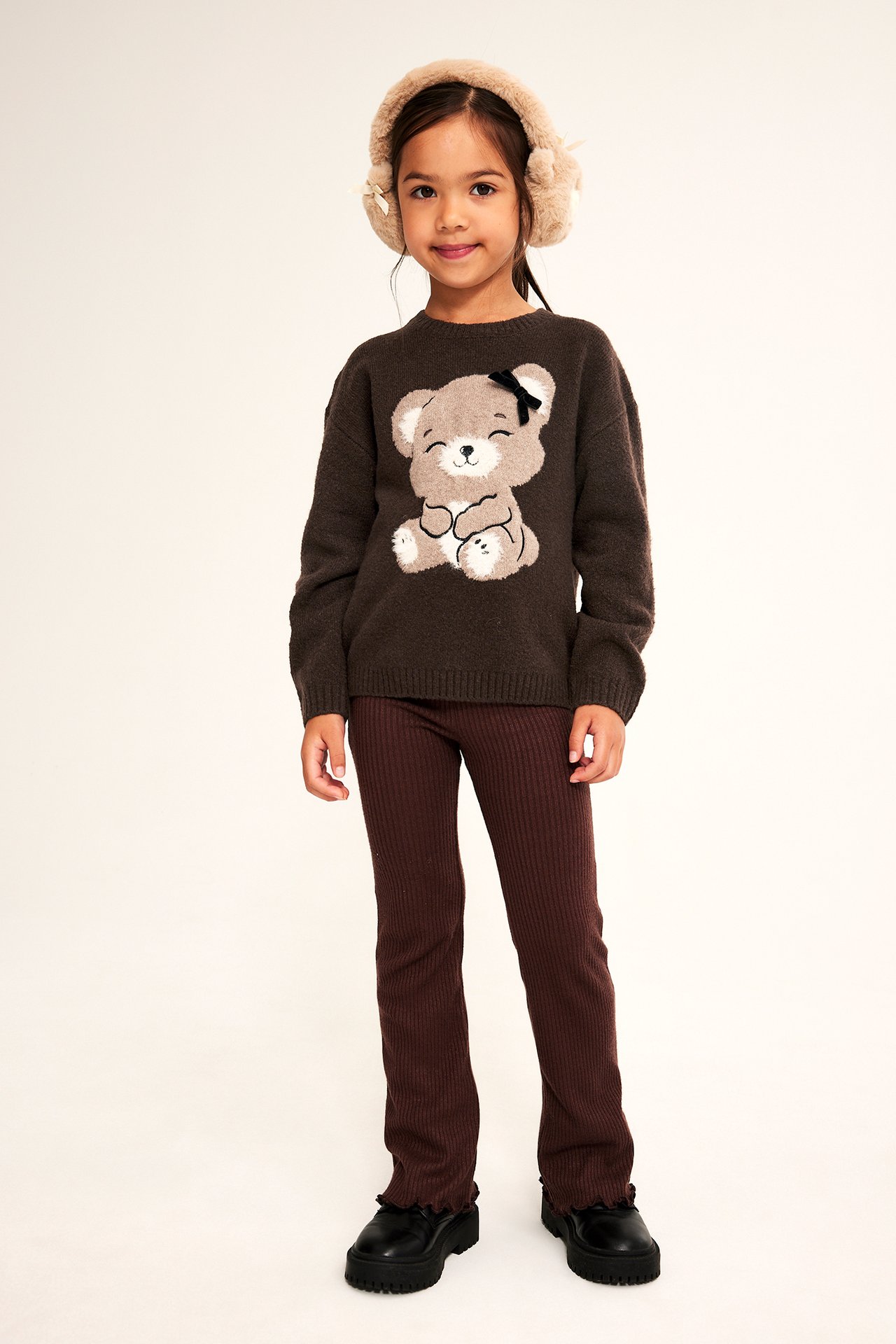 Frontbilde av et barn i brun teddybjørngenser, brune ribbestrikkede leggings, svarte sko og beige øreklokker.