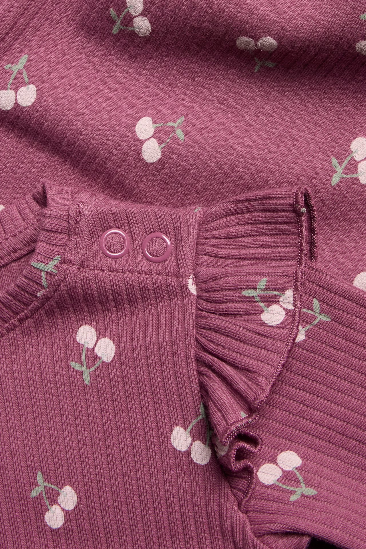 Närbild av plommonfärgad ribbad babybody med vitt körsbärstryck, axelvolanger, två tryckknappar.