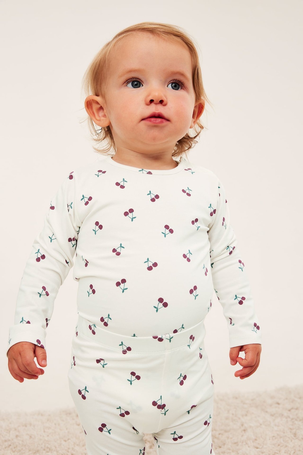Frontbilde av baby i kremfarget langermet topp og matchende leggings med kirsebærtrykk.