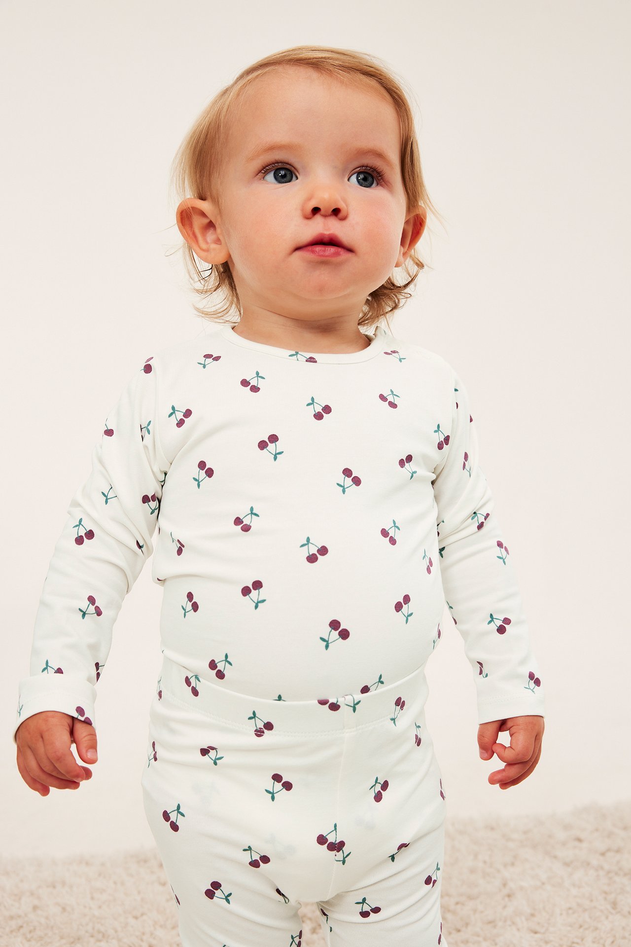 Frontvy av baby i krämfärgad långärmad topp och matchande leggings med körsbärstryck.