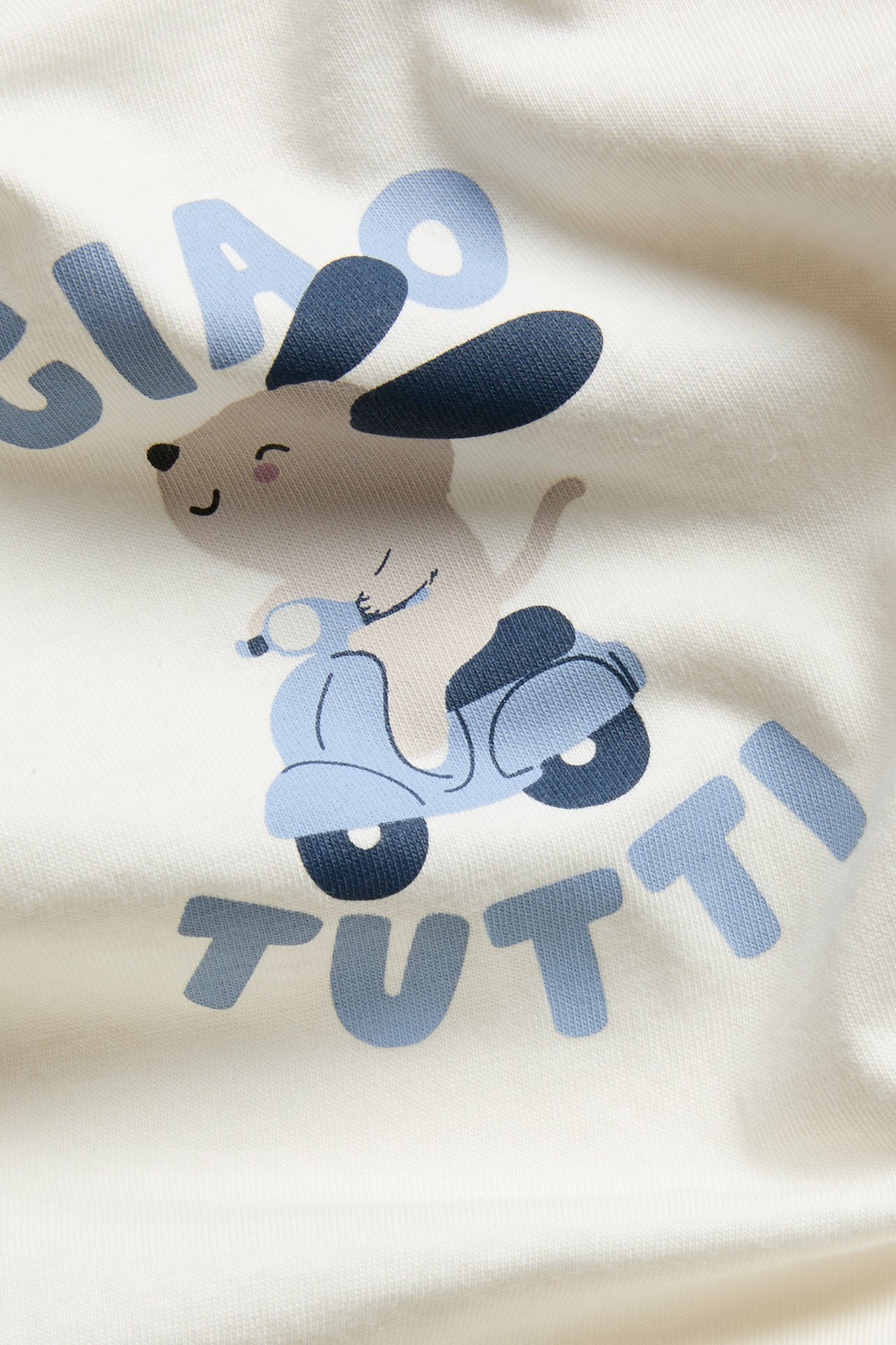 Lähikuva luonnonvalkoisesta vauvanbodysta, jossa printti: koira sinisellä skootterilla ja "CIAO TUTTI".