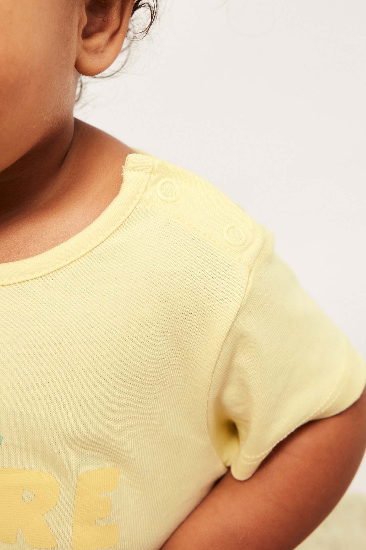 Zbliżenie dziecka w jasnożółtym body bawełnianym z nadrukiem i napami na ramieniu.