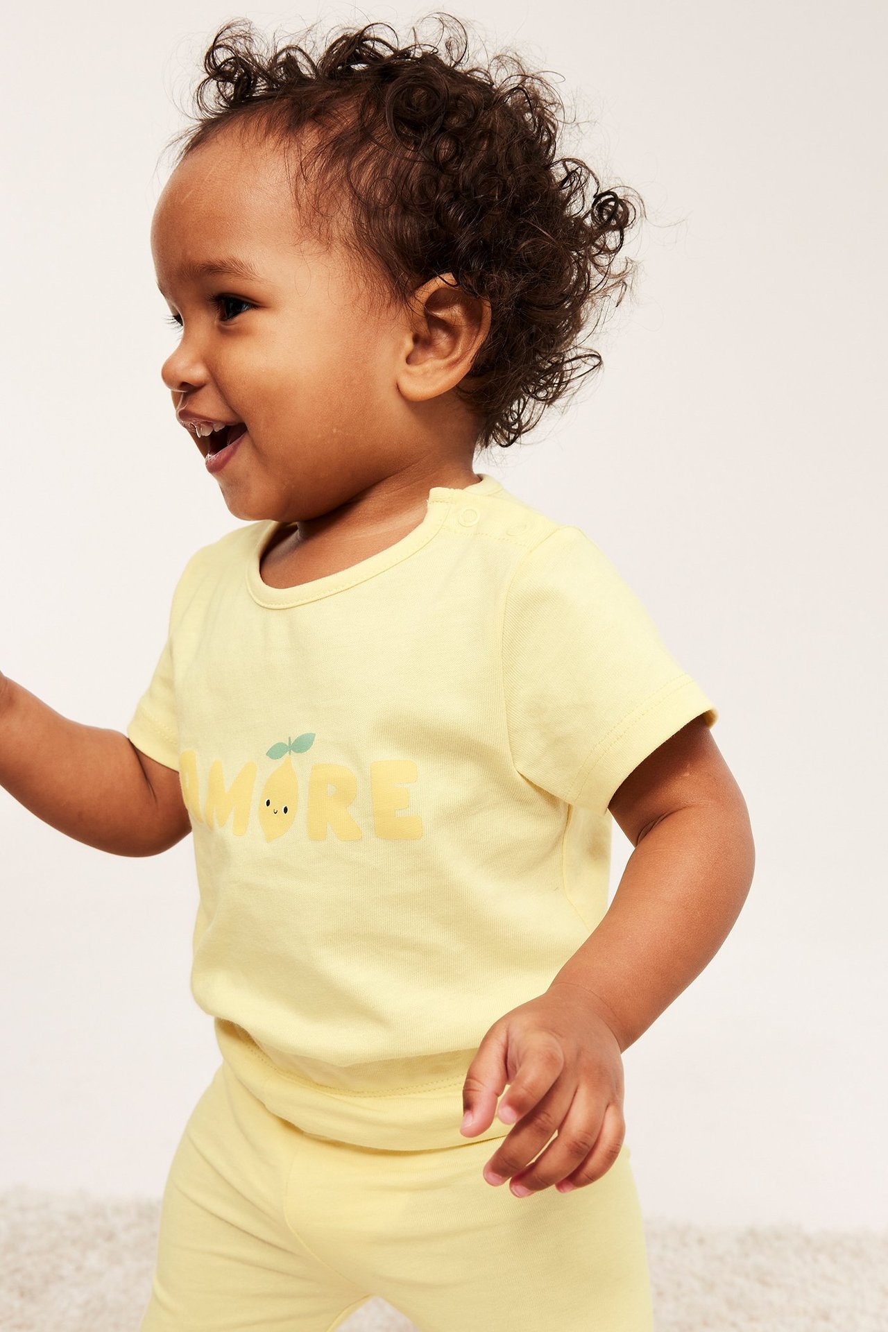 Sidovy av en glad baby i ljusgul kortärmad body med citron-print "AMORE" och matchande byxor.
