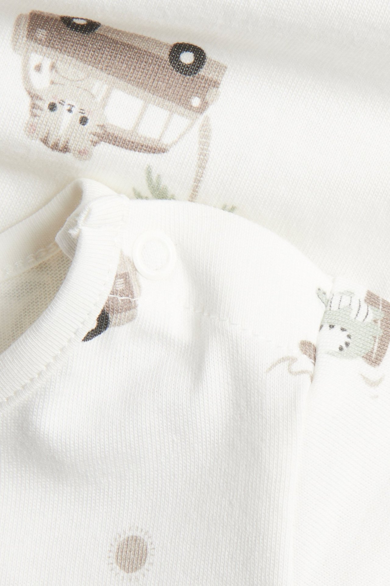 Närbild på beige bomullsbody för baby med djur- och bilmönster, samt tryckknappar på axeln.