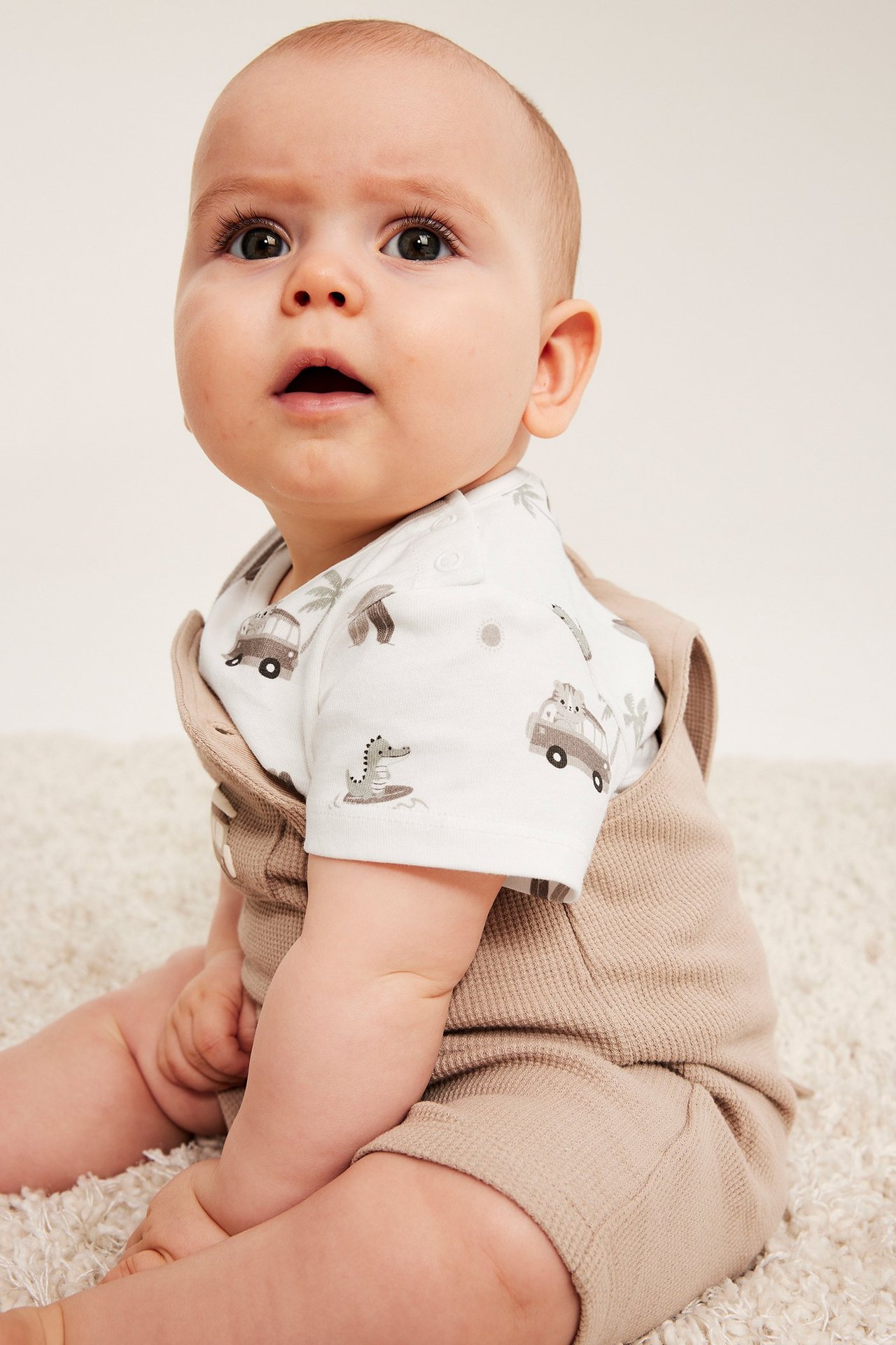 Från sidan-framifrån: baby i vit kortärmad body med djurtryck under en beige våffelstickad overall.