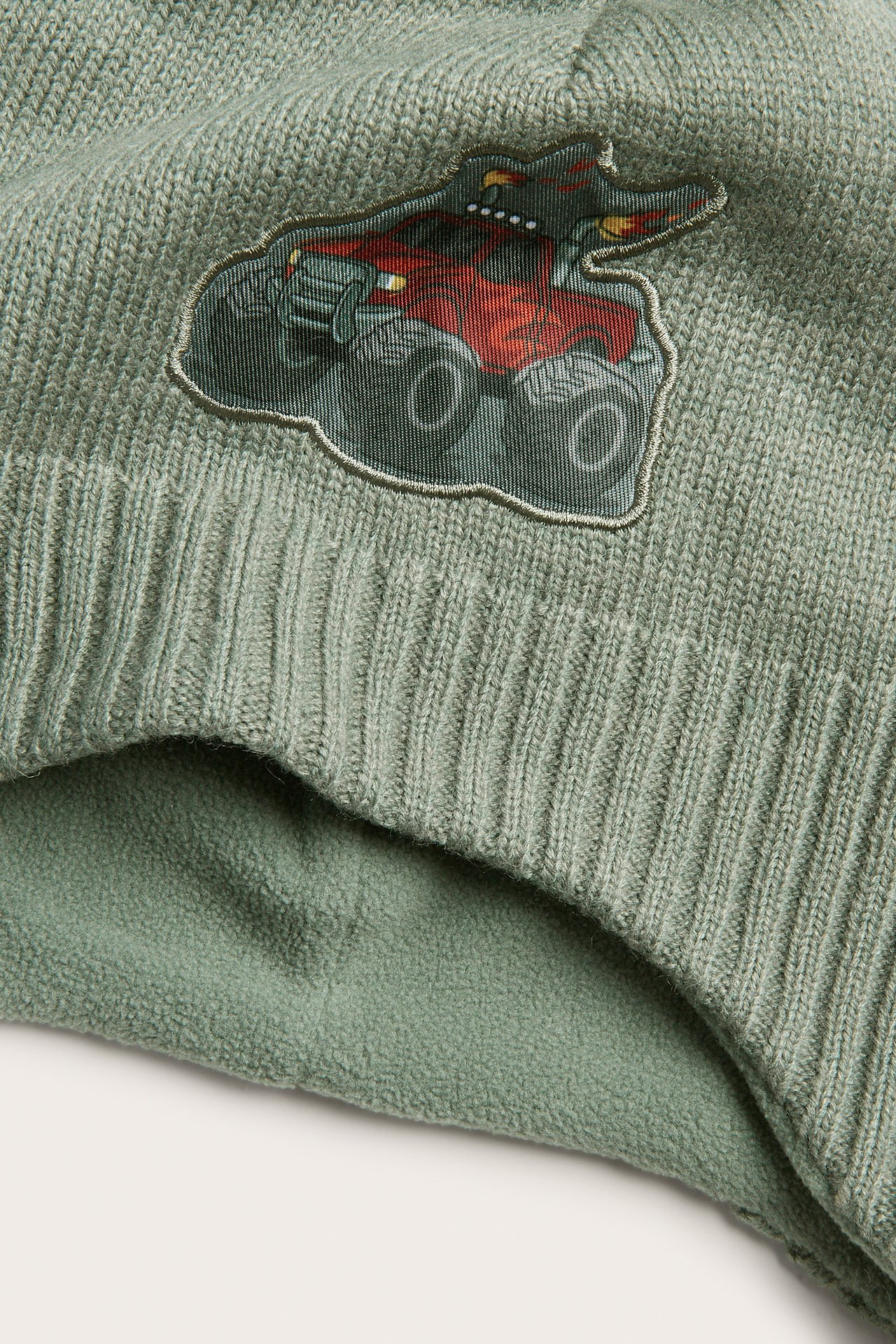 Närbild av grön stickad mössa med rött monstertruck-märke och fleecefoder.