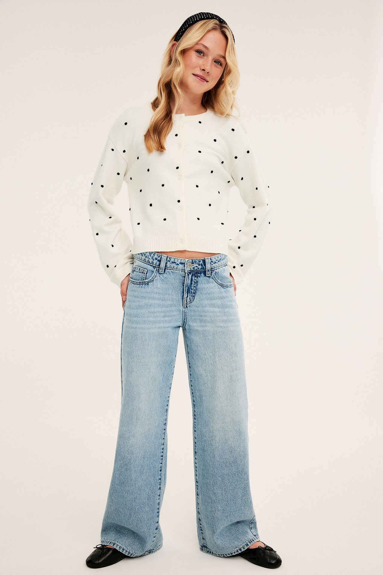 Front view: kvinne i kort kremfarget prikkete cardigan, lys blå vide jeans og svarte ballerinasko.