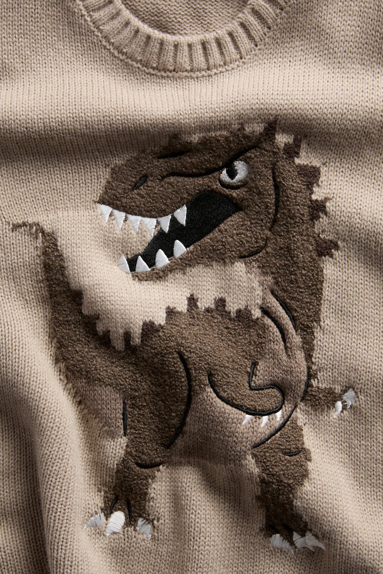 Lähikuva beigestä neulepaidasta, jossa on ruskea teksturoitu dinosaurusapplikaatio ja valkoiset hampaat.