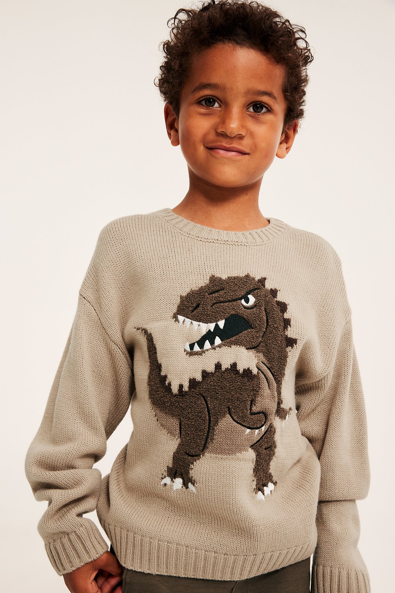 Edestä kuvattu lapsi yllään beige neulepusero, jossa ruskea dinosauruskuvio.