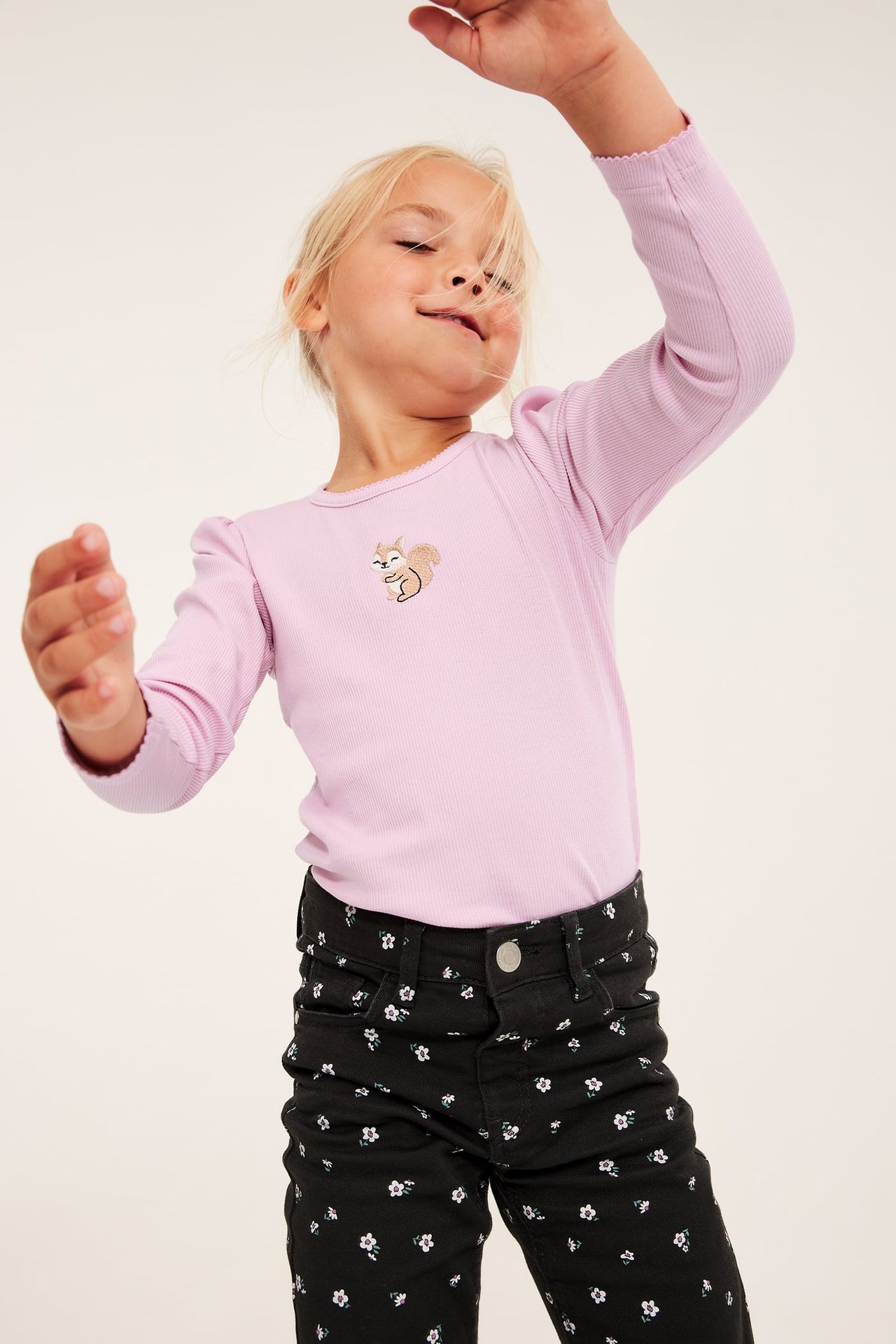 Framsida av barn i rosa ribbad långärmad topp med ekorrbroderi och svarta blommiga jeans.