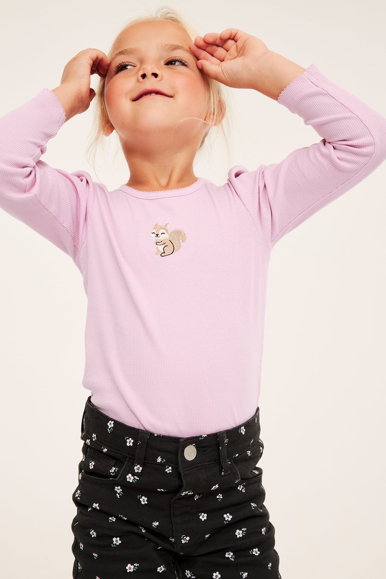 Framsida av ett barn som bär en lavendelfärgad långärmad topp med ekorrbroderi och svarta blommiga byxor.