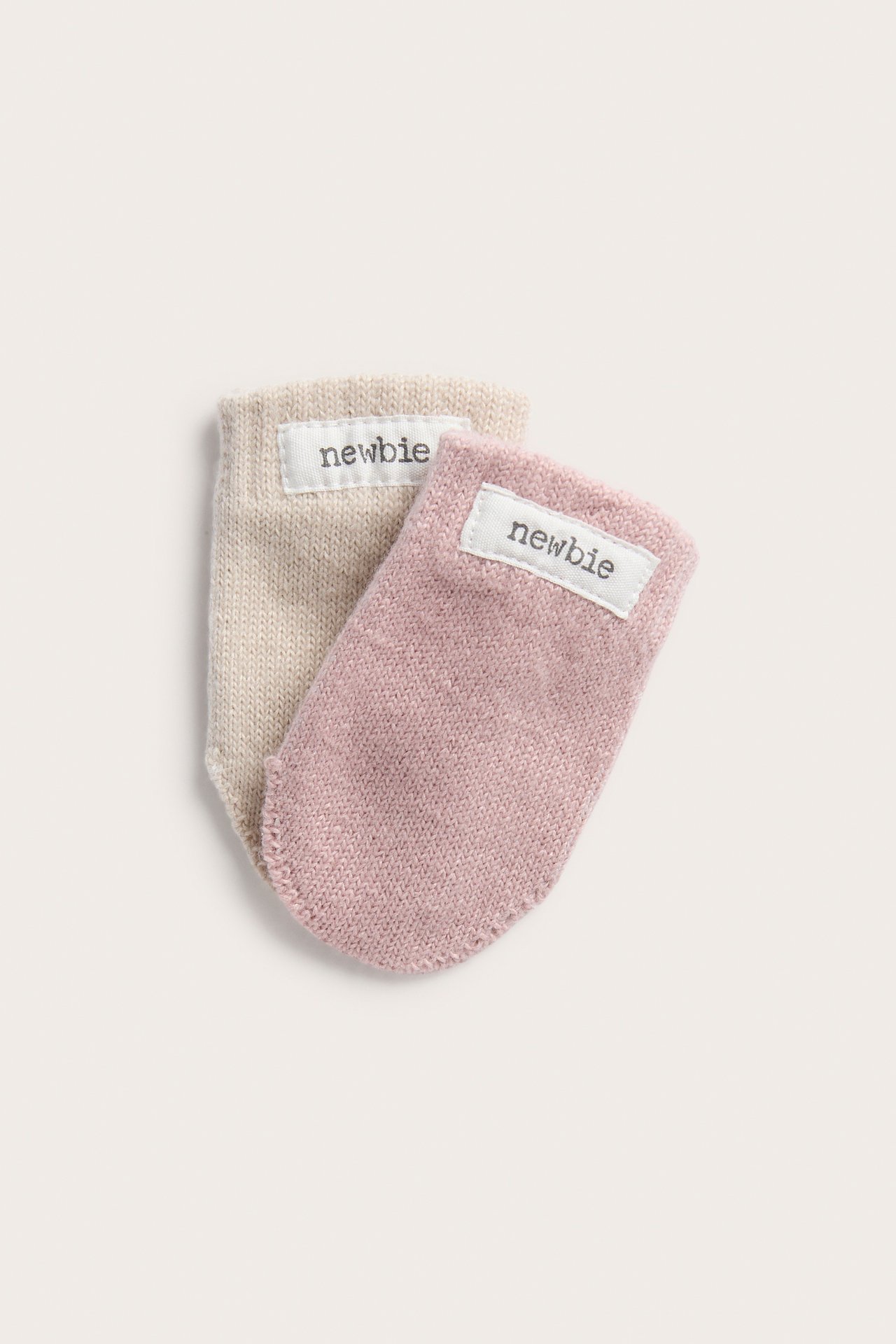 Nærbilde av to babyvotter i strikk, en rosa og en beige, med 'newbie' etiketter.