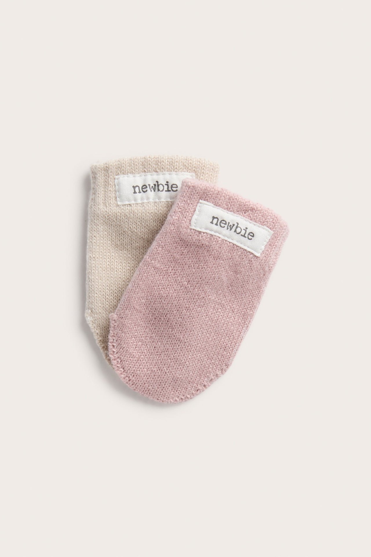 Lähikuva kahdesta neulotusta vauvan lapasesta, yksi vaaleanpunainen ja yksi beige, 'newbie'-etiketeillä.