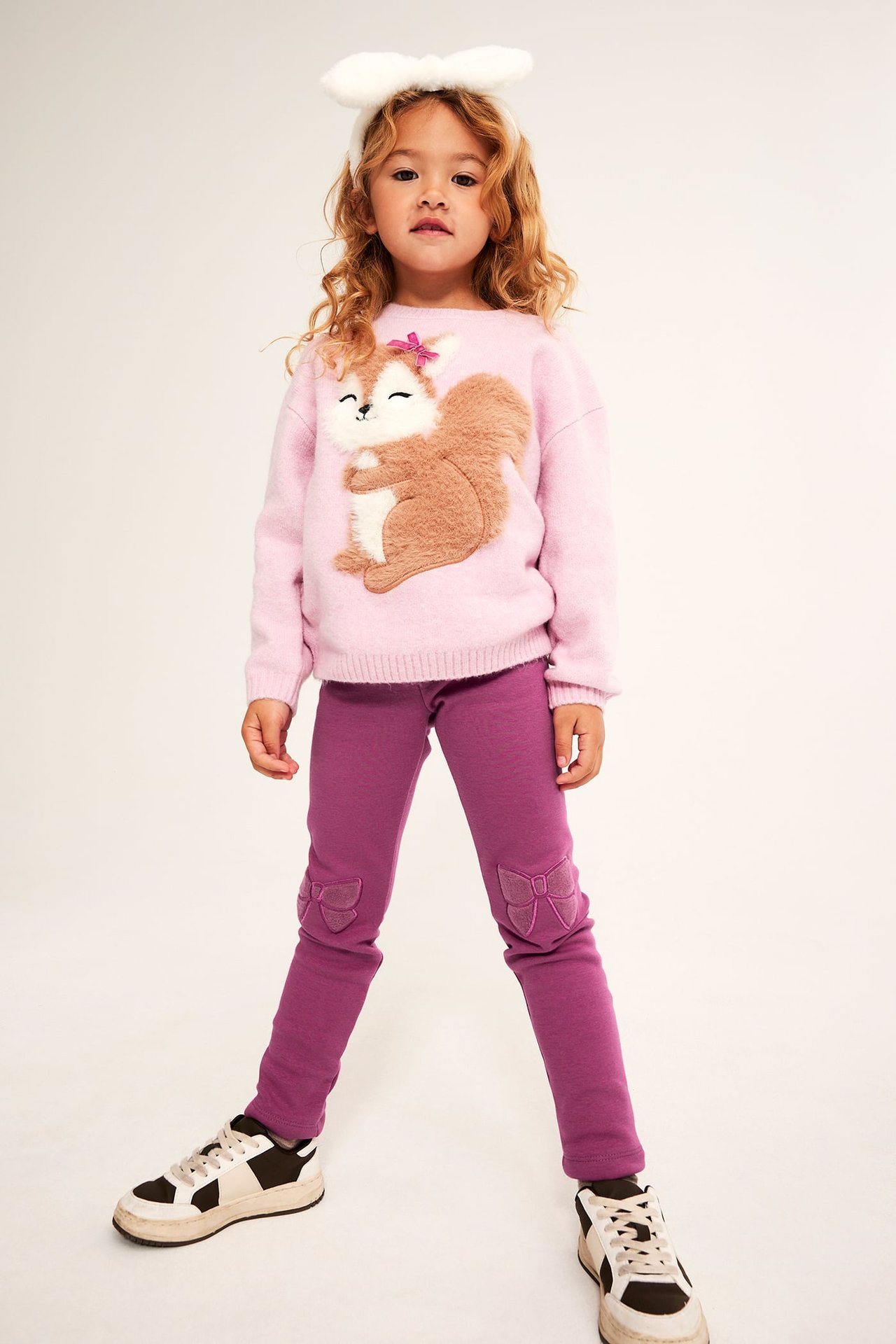 Framsida: Barn bär rosa tröja med ekorre, lila leggings med knärosetter och vitt kanin-diadem.