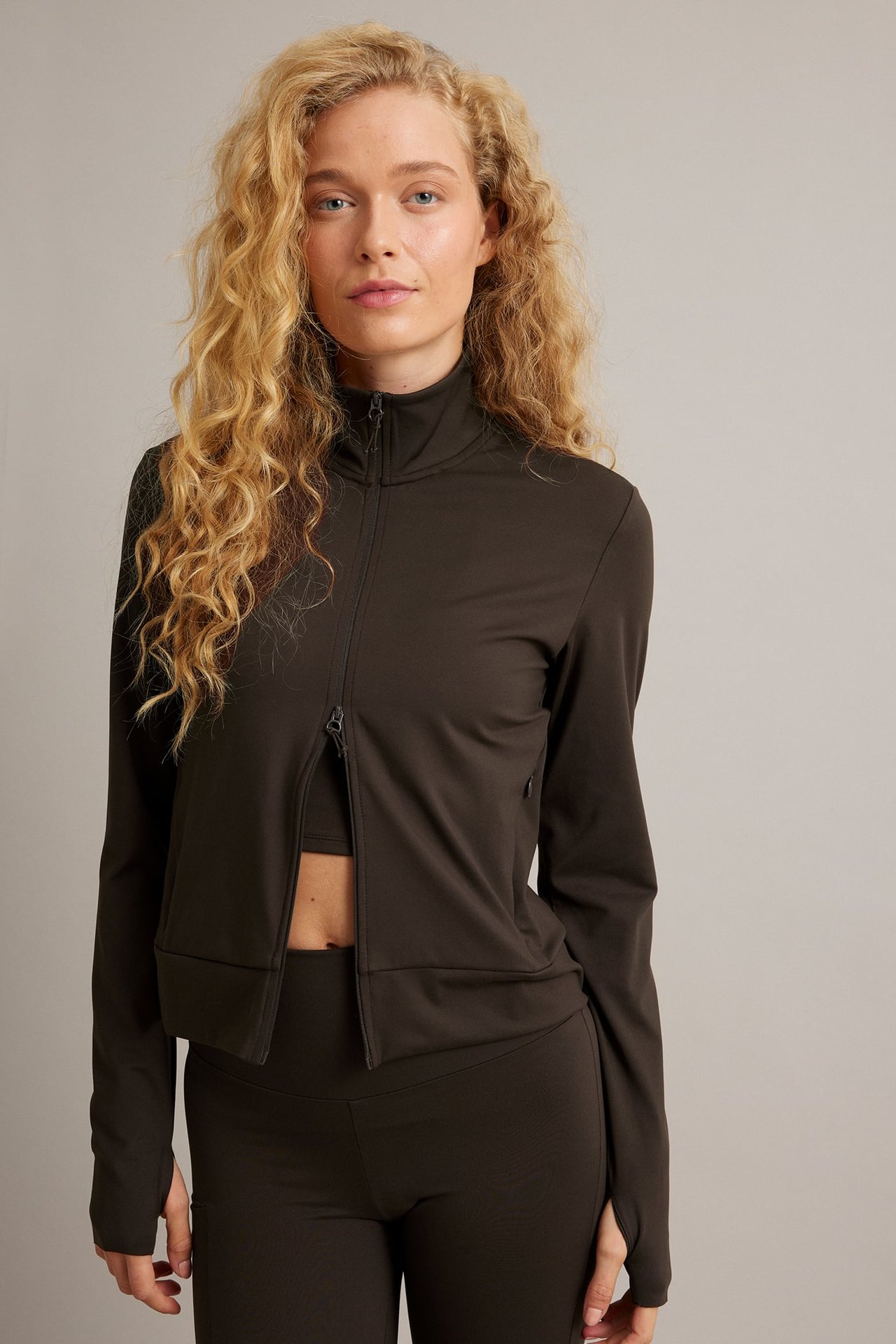 Front vy av mörkbrun långärmad jacka med dragkedja, ståkrage, crop top och matchande leggings.