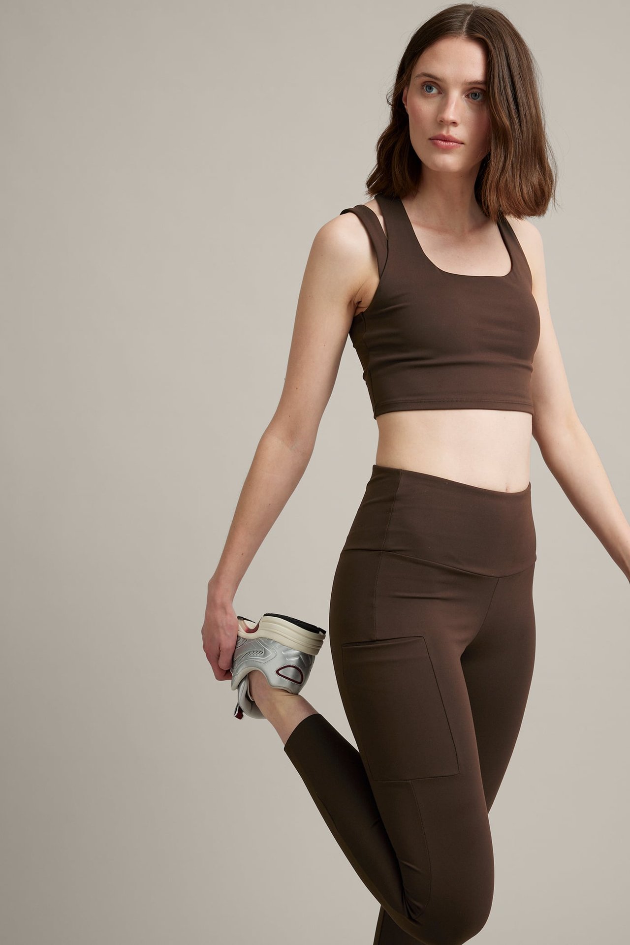 Framifrån: Kvinna i bruna träningsleggings med sidficka och brun croptop, håller silverfärgad sko.