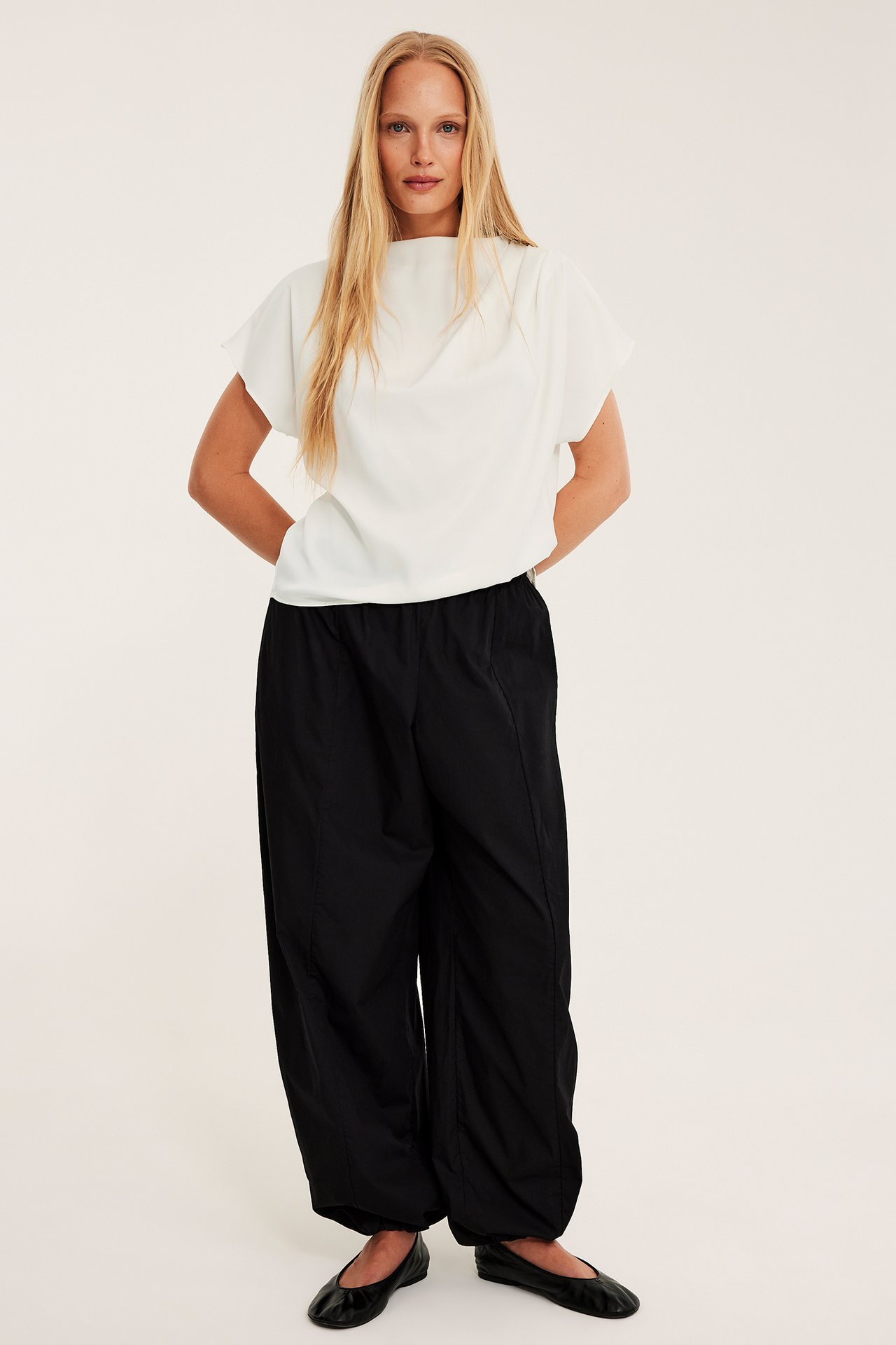 Frontvisning av en kvinne i offwhite drapert satengbluse, svarte vide bukser og svarte ballerinasko.