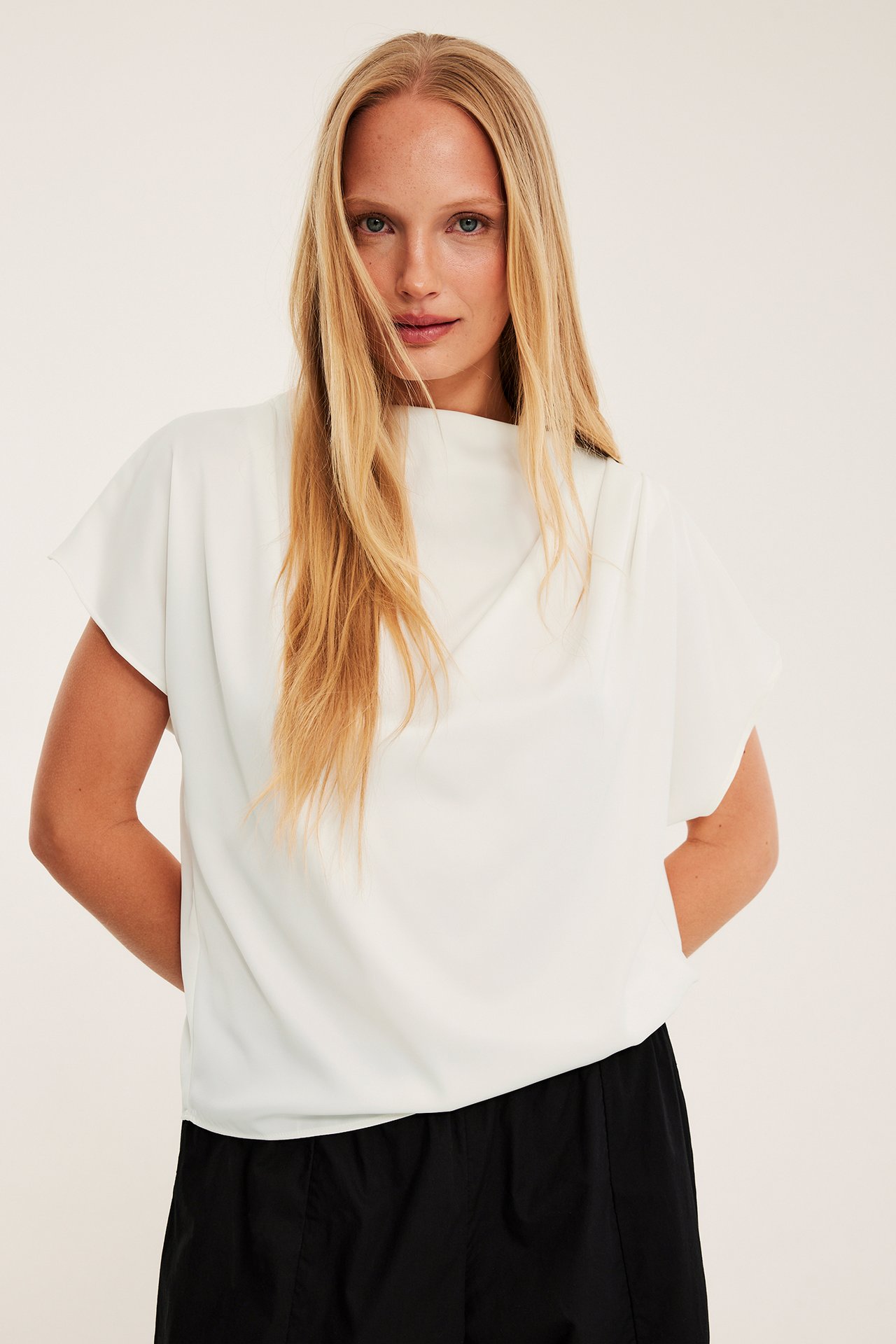 Frontbilde av kvinne i en offwhite drapert satinbluse og svarte avslappede bukser.