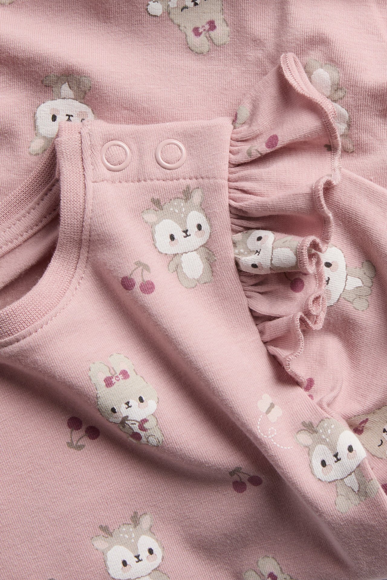 Närbild på ett rosa babyplagg med sött djur- och körsbärstryck, med tryckknappar och volanger.