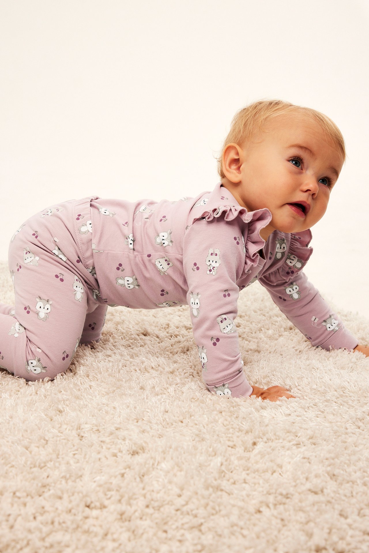 Sidebilde av baby som krabber i en lilla langermet sparkedress med kanin- og kirsebærtrykk.