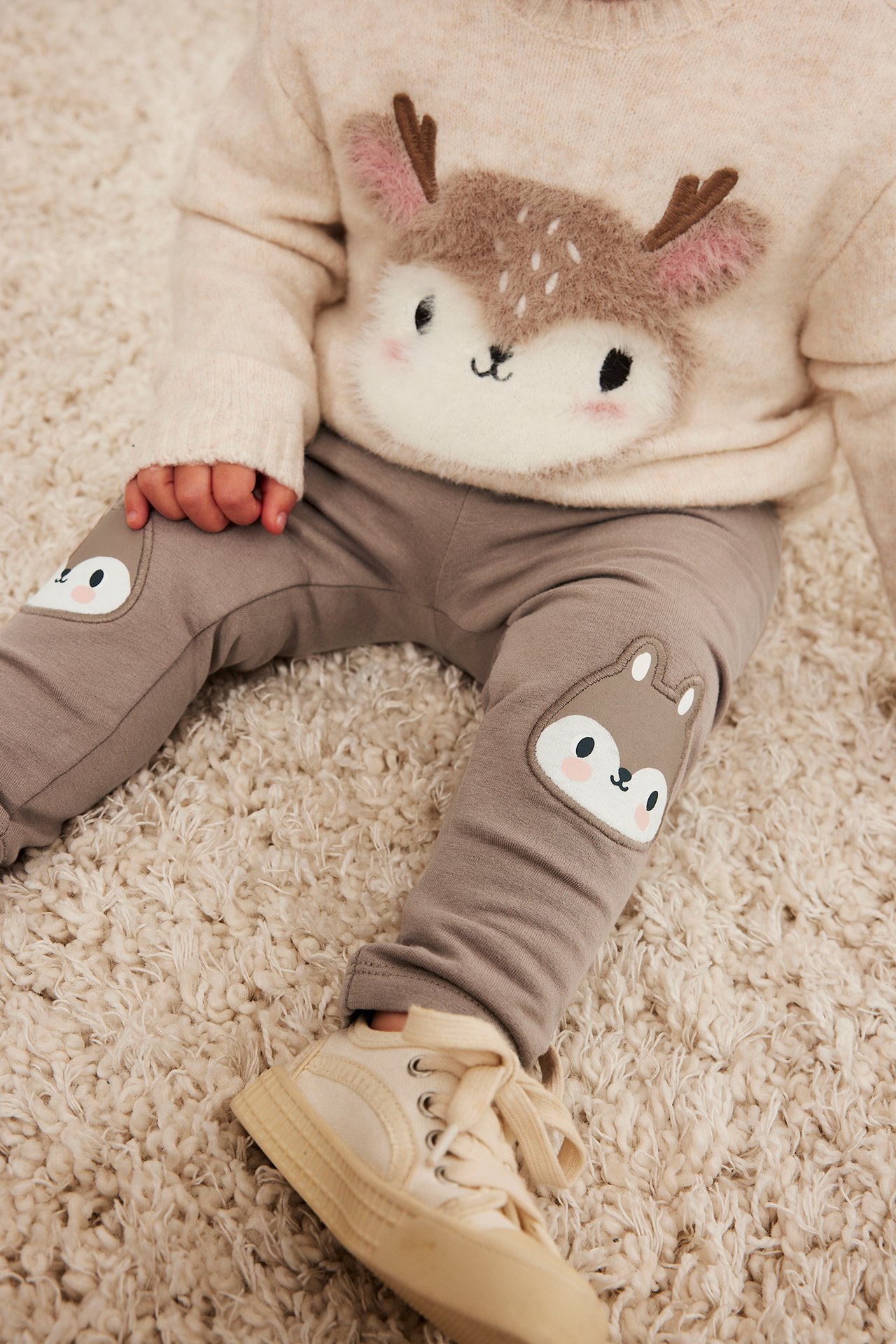 Nærbilde av en baby i beige genser med rådyr, brune leggings med dyreknelapper og kremfargede sneakers.