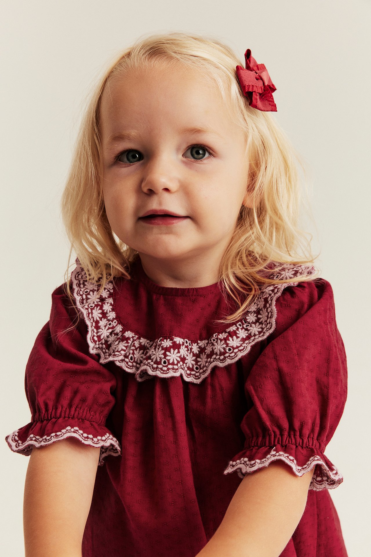 Framifrån, baby flicka i röd broderad krage klänning och röd hårrosett.