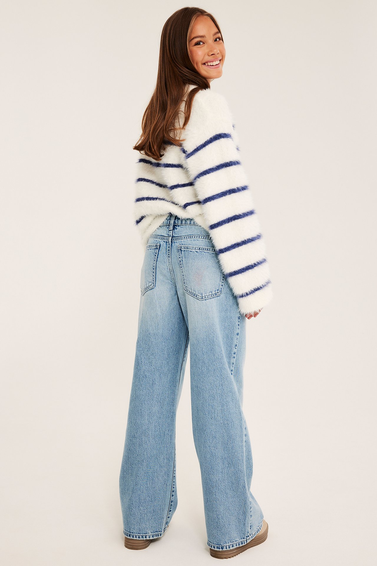 Bakfra av et barn som har på seg en hvit luftig stripet genser med lyseblå vide jeans.