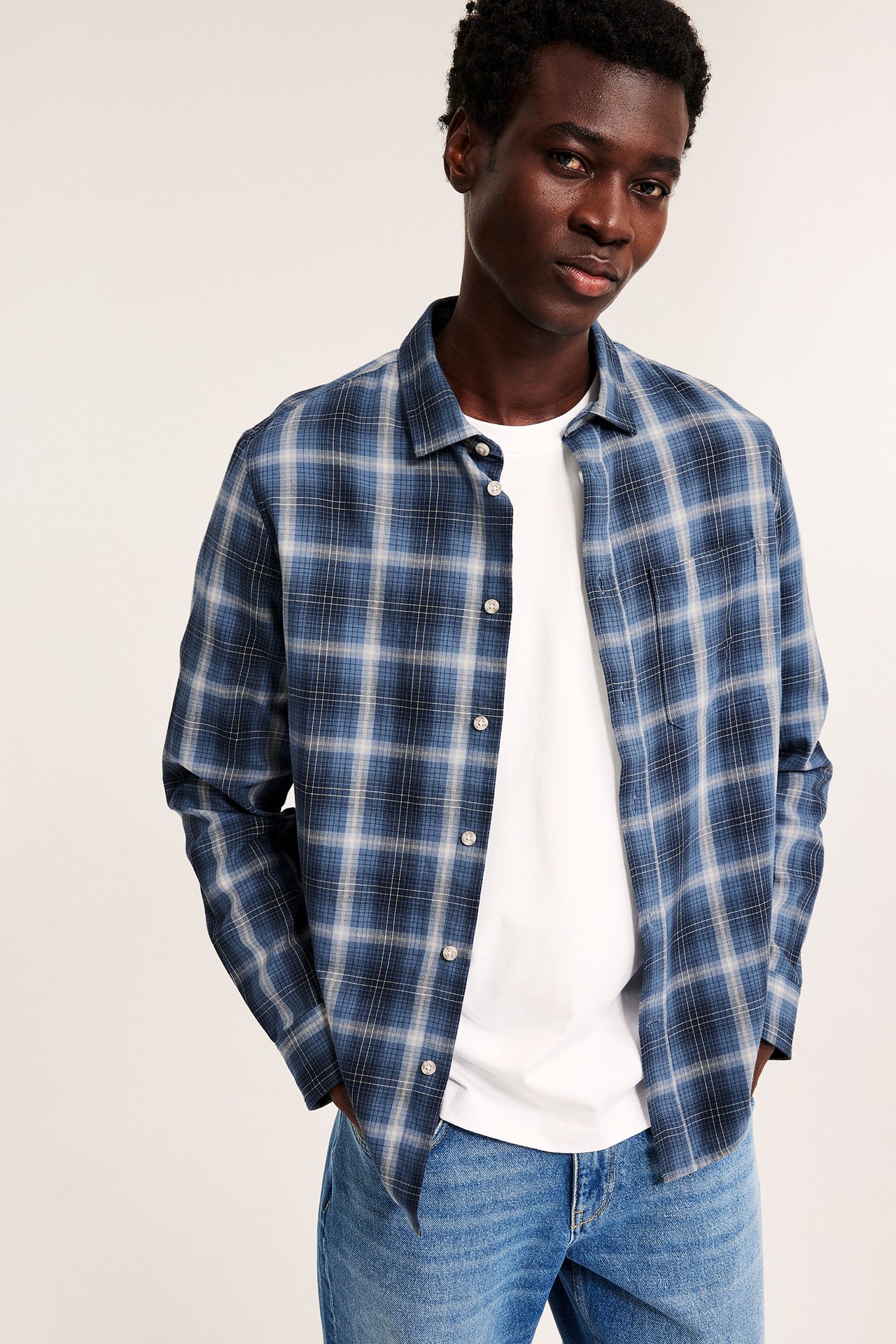 Frontbilde av mannlig modell iført blå rutete langermet skjorte over hvit t-skjorte og blå jeans.