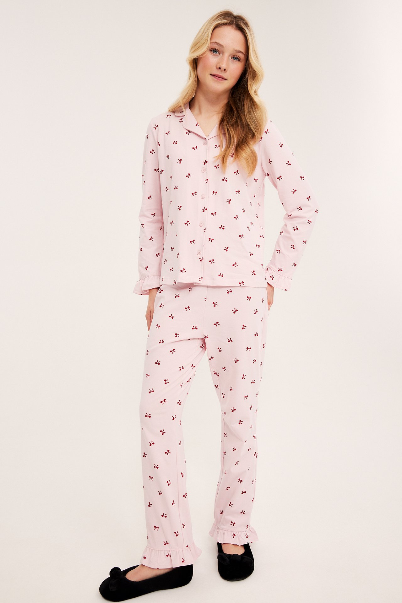 Framifrån: kvinnlig modell i rosa pyjamas med rött rosettmönster och svarta tofflor.