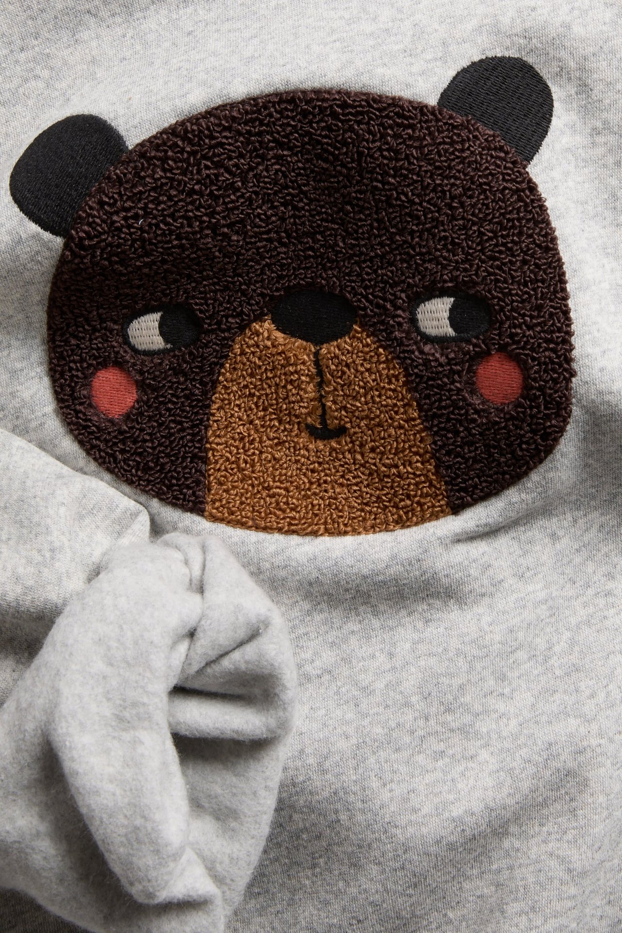 Nærbilde av en grå sweatshirt med teksturert brun bjørneansiktapplikasjon.