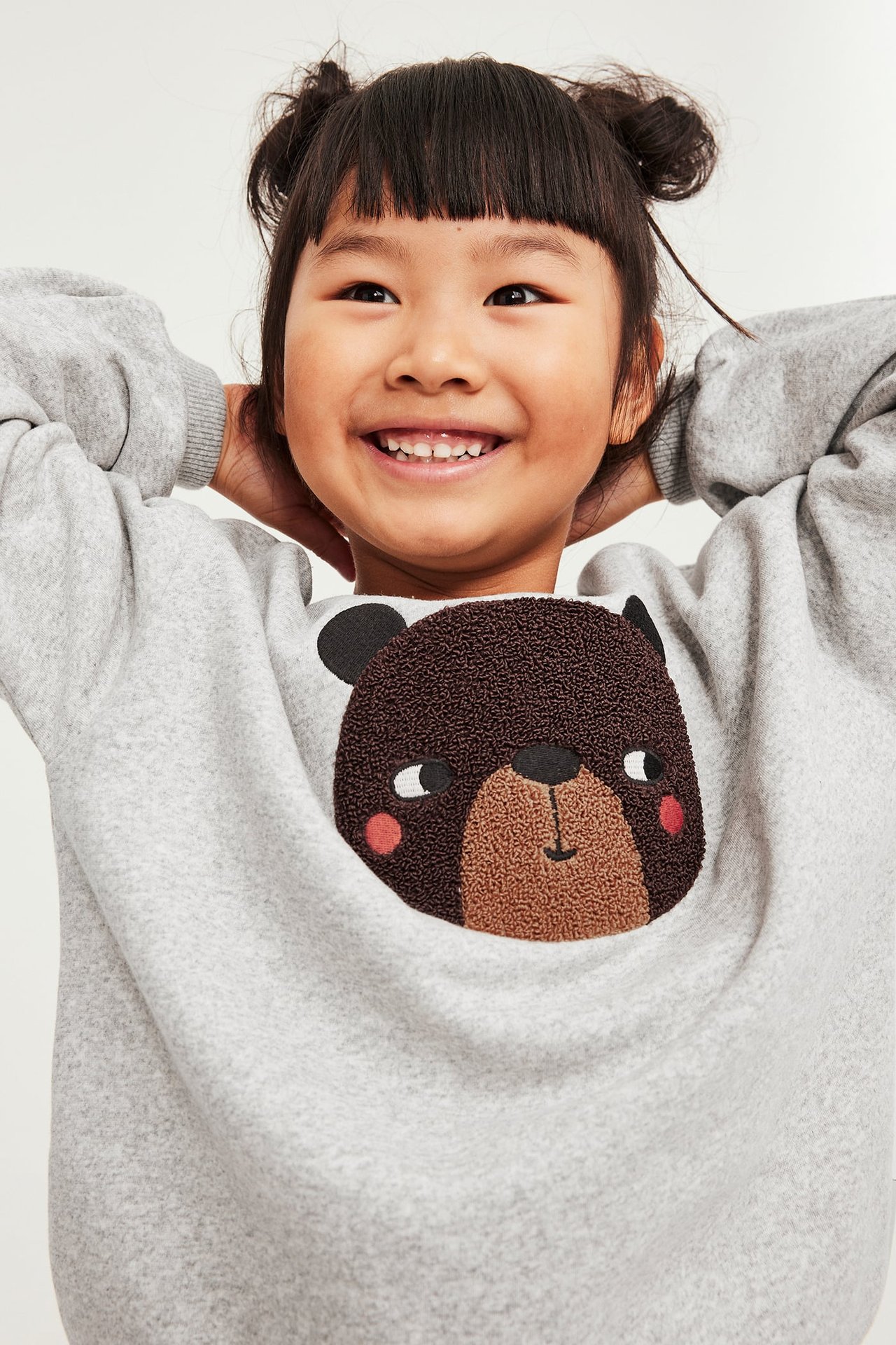 Forfra, barn i en lys grå sweatshirt med et brunt brodert bjørneansikt.