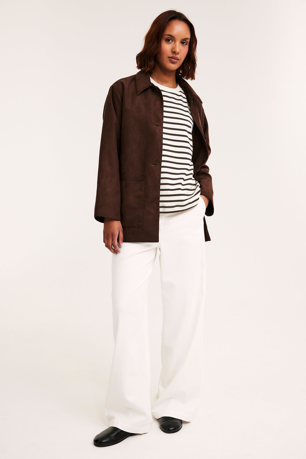 Framifrån: Kvinna i oversized vit och mörkbrun randig t-shirt, brun jacka, vita byxor, svarta skor.