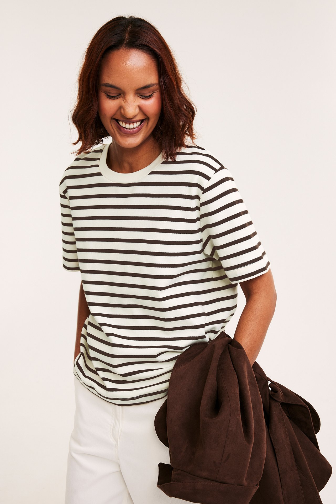 Leende kvinna, framifrån, i randig oversized t-shirt i benvitt och mörkbrunt, vita byxor, håller i en brun jacka.