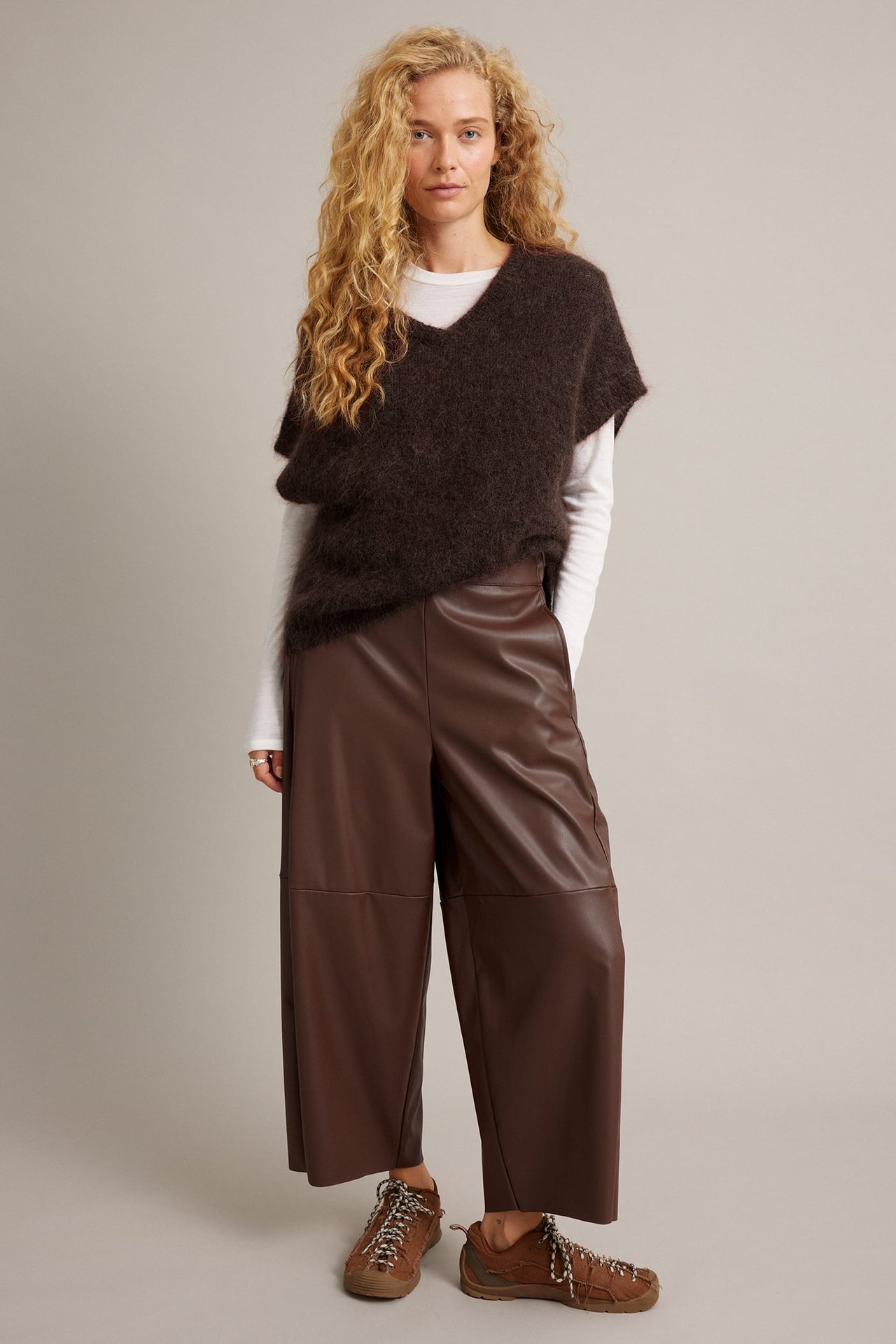 Front view: kvinne i brune cropped bukser i imitert skinn, mørkbrun lodden genser, hvit topp, brune joggesko.