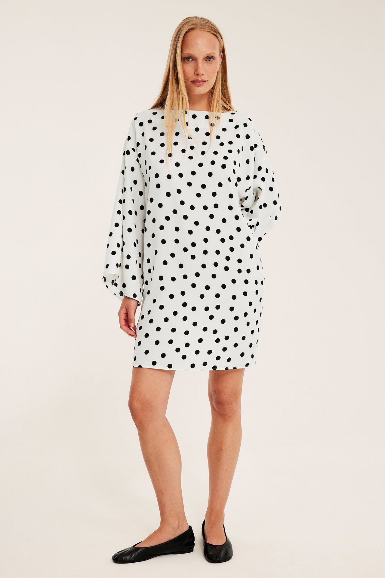 Nainen lyhyessä luonnonvalkoisessa pilkkusatiinimekossa leveillä hihoilla ja mustilla balleriinoilla, edestä.