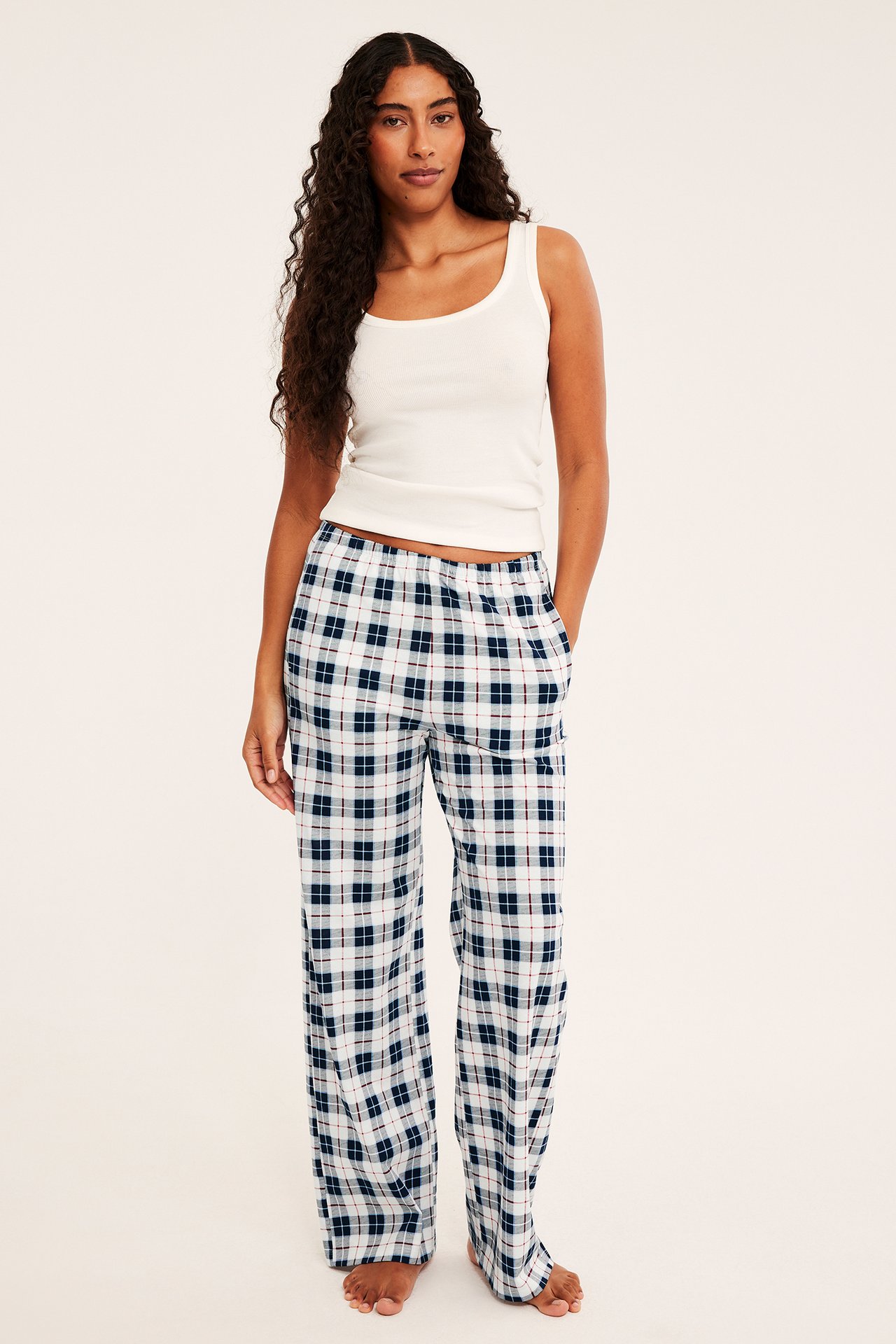 Frontbilde av voksen kvinne i hvite og marineblå rutete pyjamasbukser og en hvit ribbet singlet.