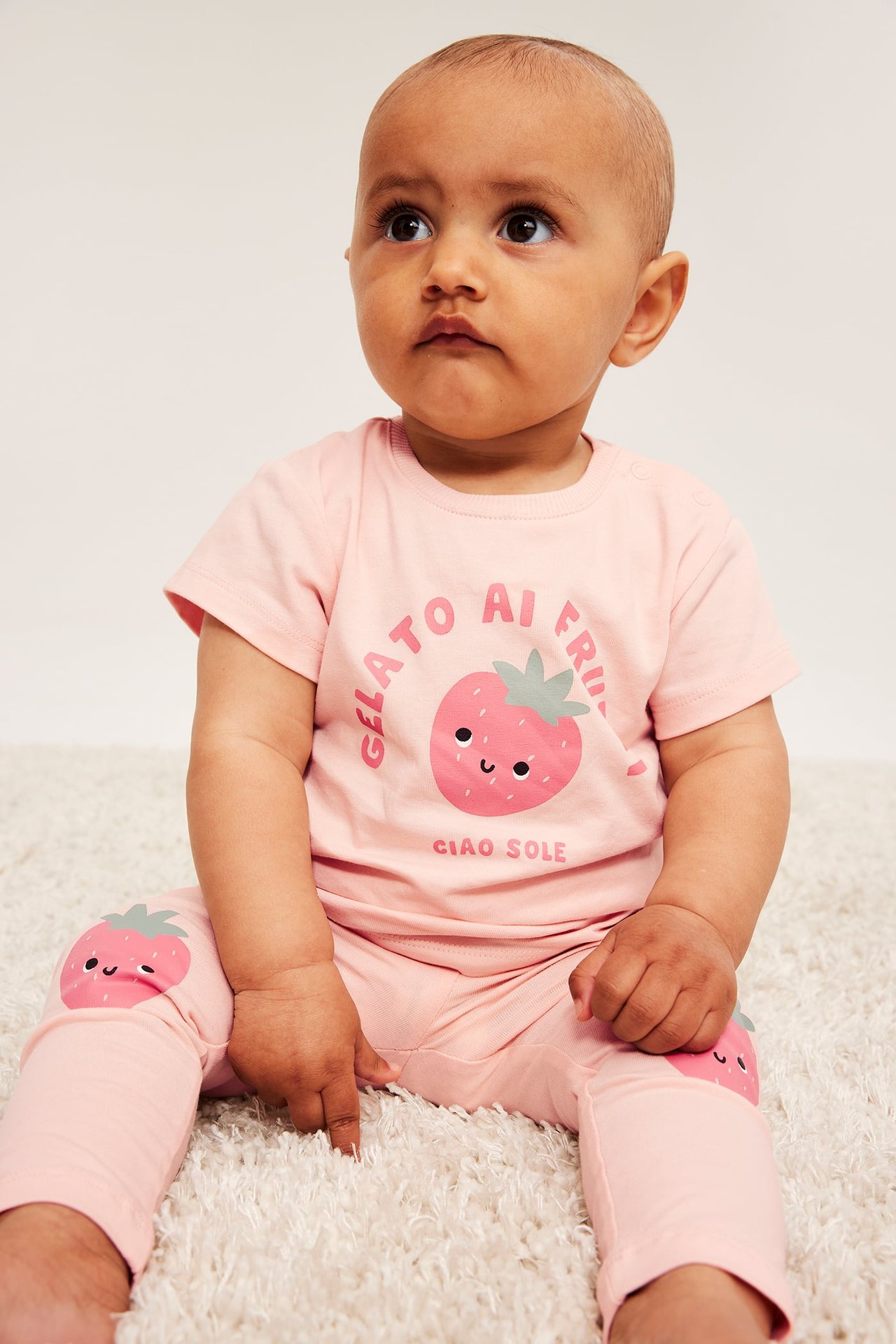 Framifrån: baby bär rosa t-shirt med jordgubbstryck, axelknappar och matchande leggings.