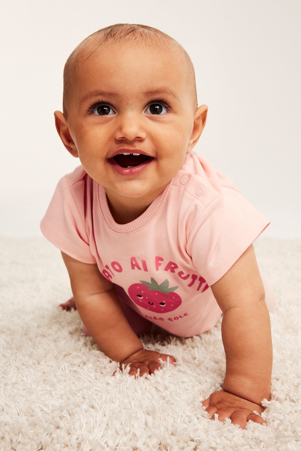 Framifrån: baby i en ljusrosa kortärmad t-shirt med jordgubbstryck och tryckknappar på axeln.
