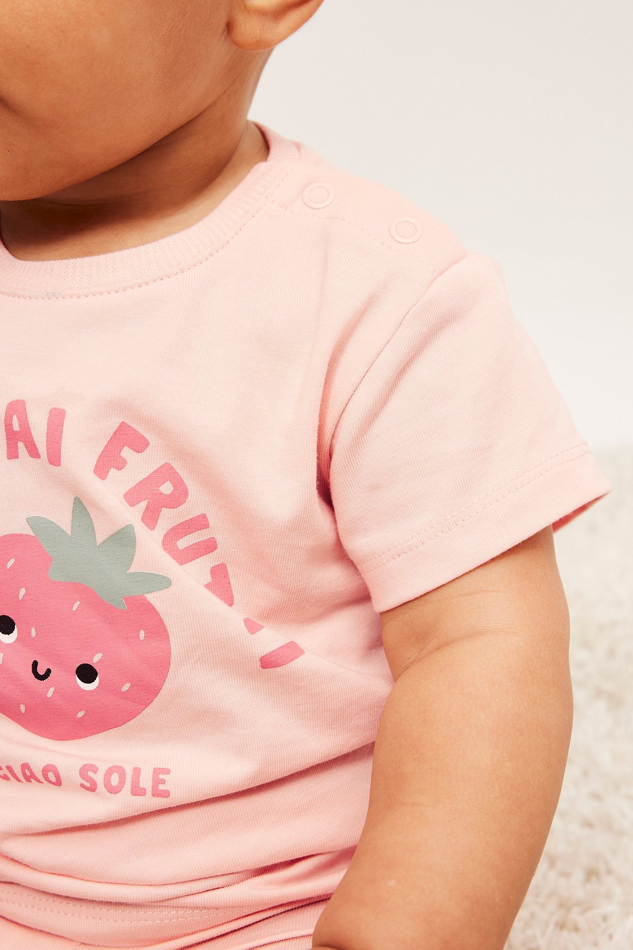 Närbild på en baby i en ljusrosa kortärmad bomulls t-shirt med tryckknappar på axeln och jordgubbstryck.